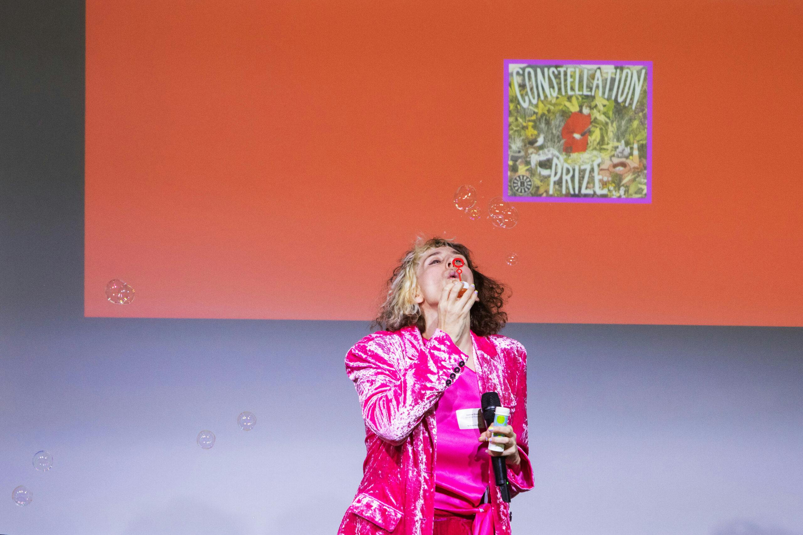 Jeanne Drach pustet in einem pinken Blazer Seifenblasen in die Luft. Im Hintergrund ist eine Orangene Slide einer Powerpoint-Präsentation zu sehen, mit dem Cover des Podcasts „Constellation Prize“.