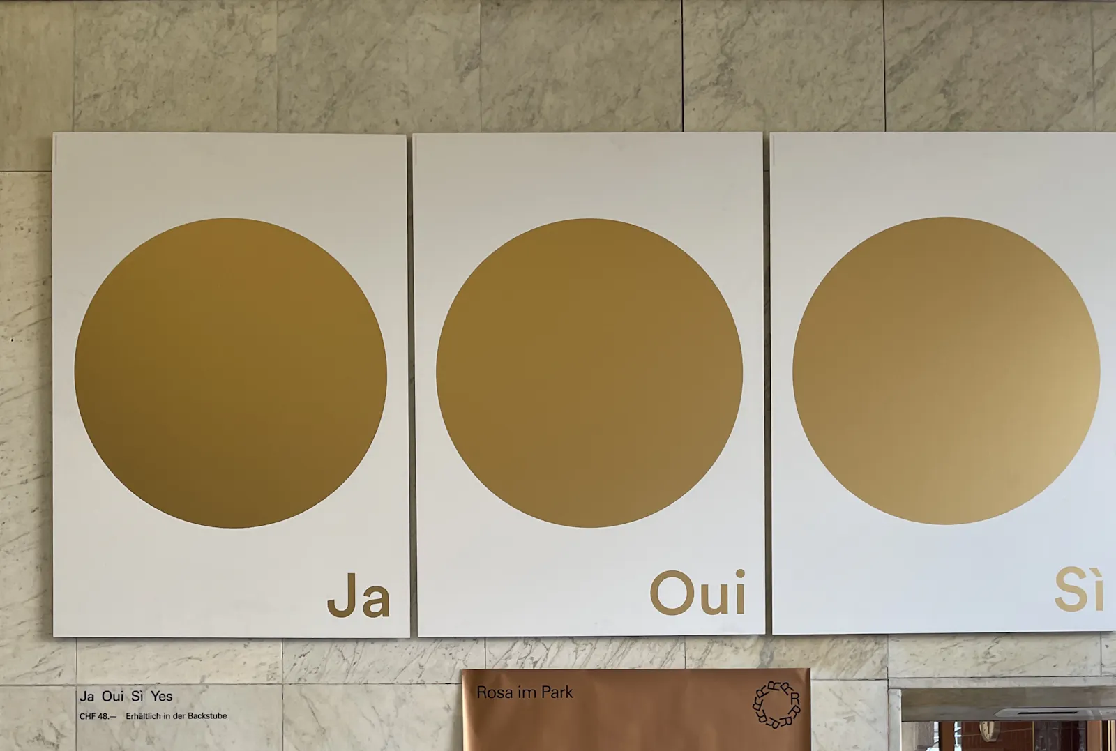 Wandinstallation: auf drei gleich große weiße Plakate sind goldene Punkte gedruckt. Jeweils rechts unten steht Ja, Oui, Sí.