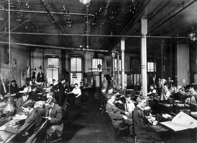The New Orleans Item newsroom, circa 1900. Schwarz und weiß bild eines sehr großen, sehr hohen Raumes. An etwa zwanzig Tischen sitzen Menschen, lesen und schreiben.