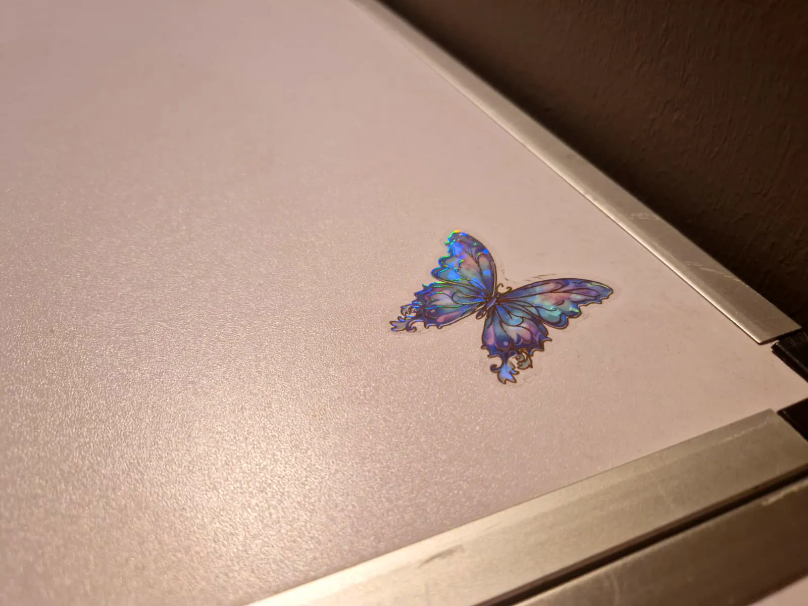 Ein glänzender, holografischer Schmetterlingsaufkleber in Blau- und Lilatönen klebt auf der hellen, strukturierten Oberfläche des DA-Tapeziertisches.