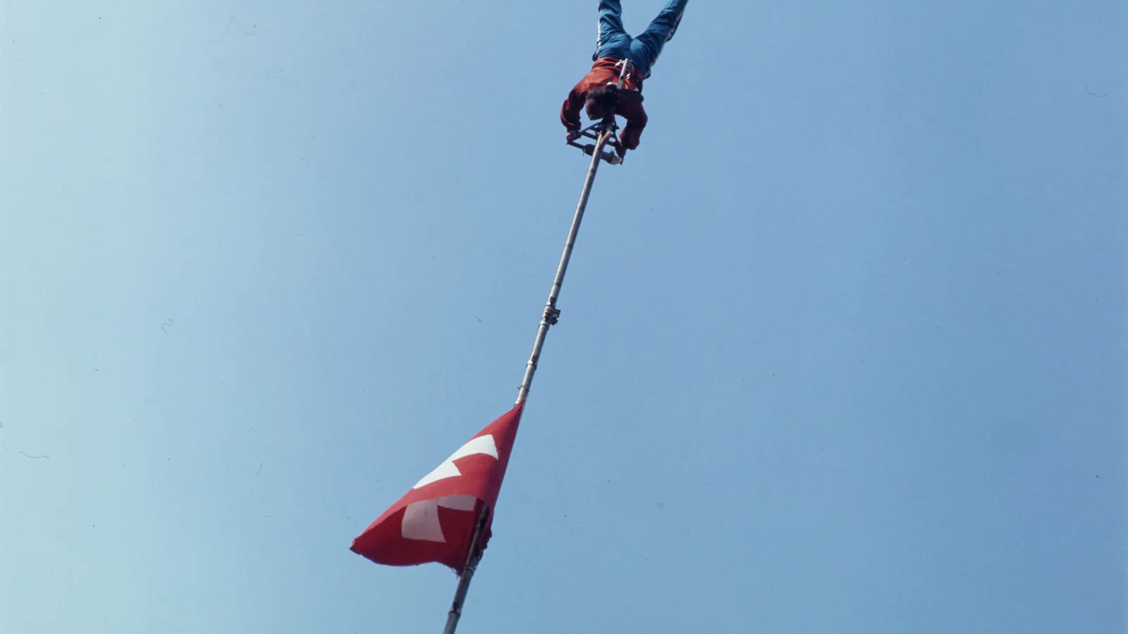 Person hängt kopfüber an der Spitze eines hohen Fahnenmasts mit Schweizer Fahne vor blauem Himmel.