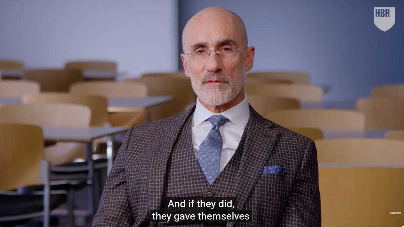 Screenshot aus dem Video von Arthur Brooks, man sieht den Professor im karierten Anzug mit blauer Krawatte in einem Hörsaal sitzen