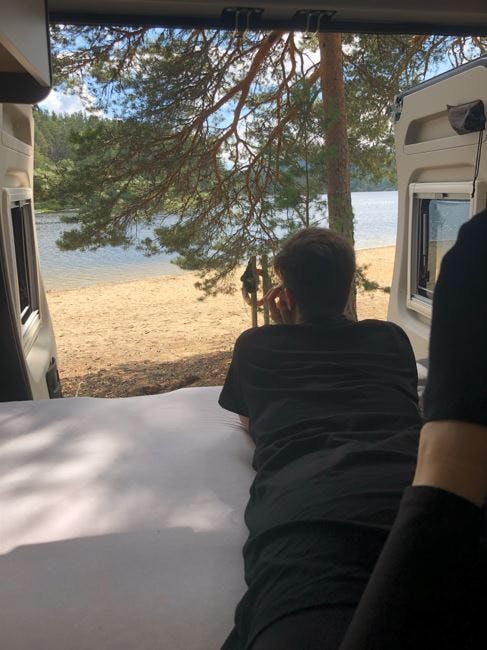 Dunkel angezogene Person liegt im Wohnmobil auf dem Bett bei geöffneter Heckklappe und schaut auf einen See