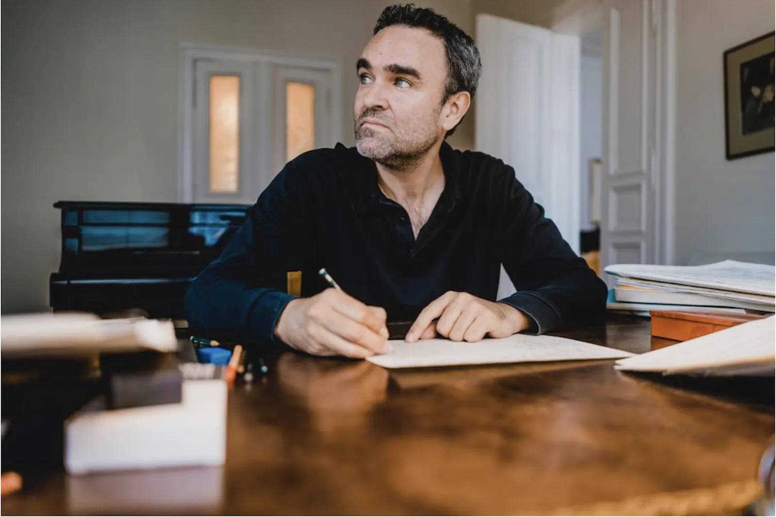 Widmann sitzt an einem Schreibtisch mit einem Bleistift in der Hand, offenbar beim Verfassen einer Partitur, und blickt nach links aus dem Bild. Im Hintergrund ein Klavier.