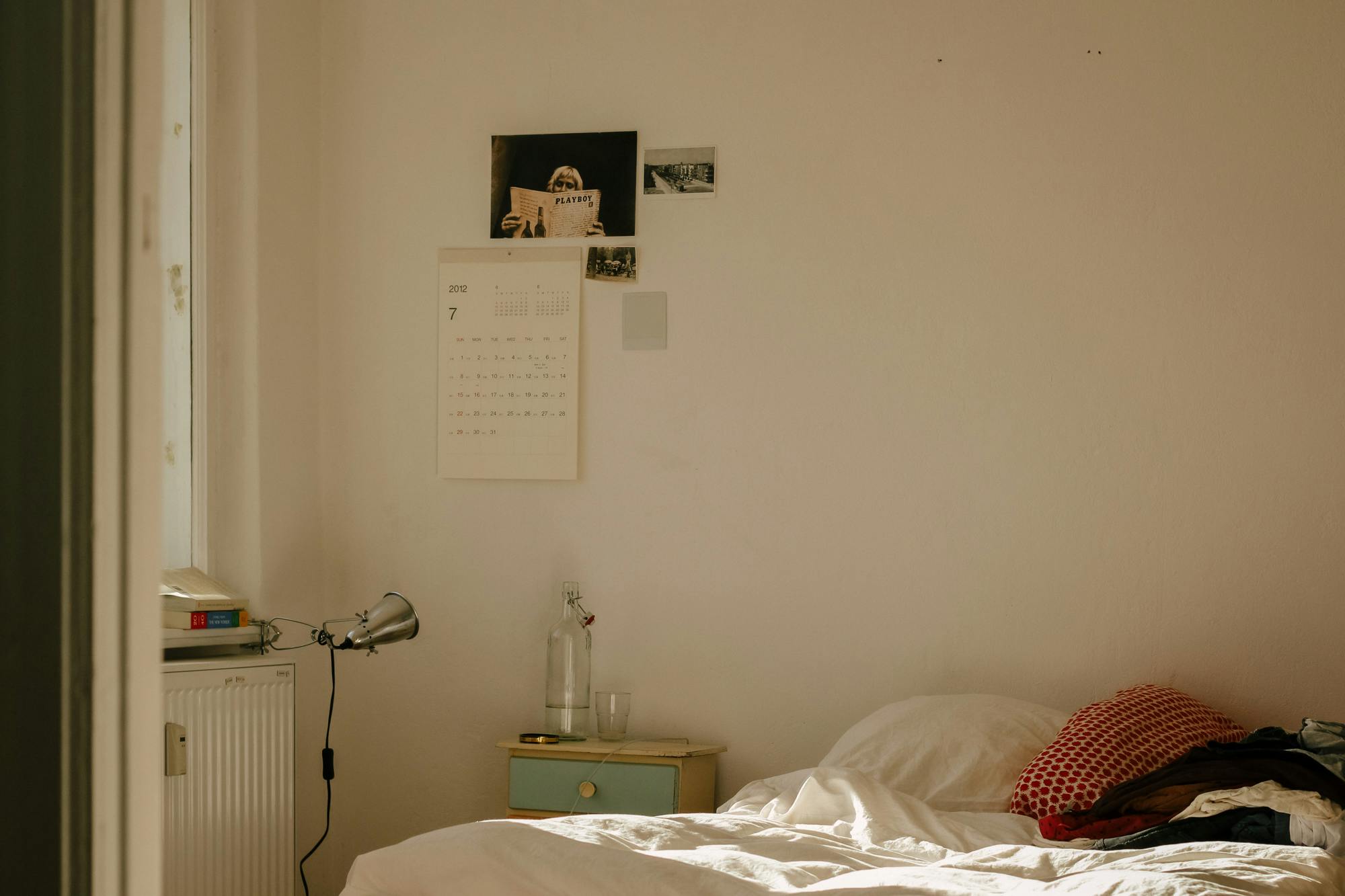 Schlichtes Schlafzimmer mit ungemachtem Bett, Leselampe und Fotografien an der Wand.