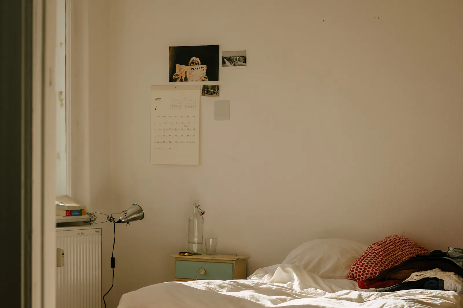 Schlichtes Schlafzimmer mit ungemachtem Bett, Leselampe und Fotografien an der Wand.