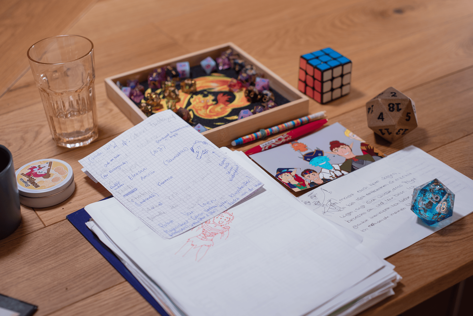 Blick auf Rubis Spieliplatz. Im Vordergrund handgeschriebene Notizen und eine Kritzelei von Jeri in ihrem neuen Outfit in roter Farbe. Darauf liegt ein kariertes Notizblatt mit weiteren Spielnotizen, u.a. zu Paddys Vater, Eleasaa und Amorilie. Rechts daneben eine Illustration der Hüter der Obelisken sowie ein blauer Liquidcore-W20 und weitere handgeschriebene Notizen. Im Hintergrund ein Fridolin Dicetray mit vielen verschiedenen Würfeln, ein 3x3 Rubik's Cube, ein faustgroßer Holz-W20, ein Trinkglas und eine Dose von Elaynies Honigbonbons.