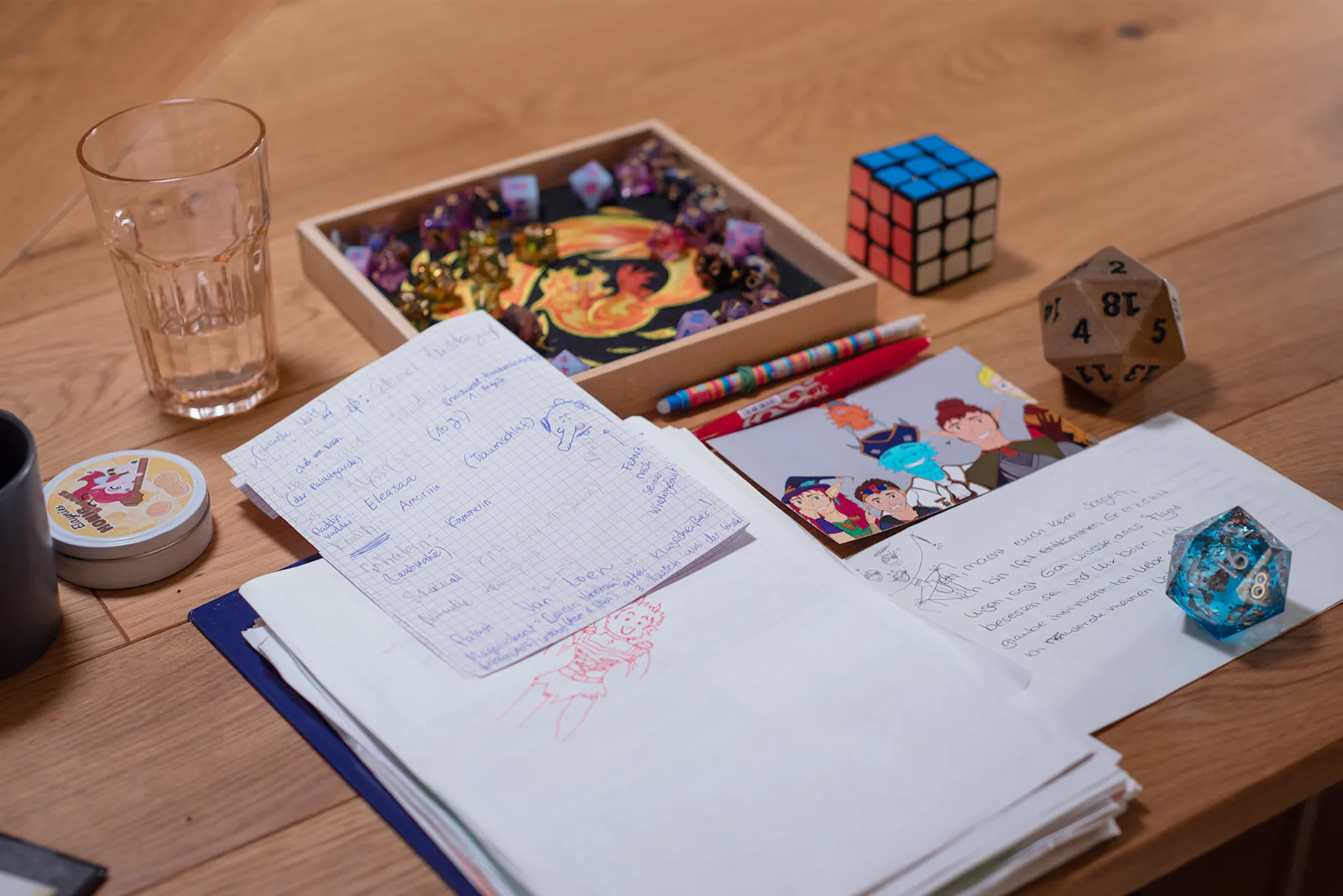 Blick auf Rubis Spieliplatz. Im Vordergrund handgeschriebene Notizen und eine Kritzelei von Jeri in ihrem neuen Outfit in roter Farbe. Darauf liegt ein kariertes Notizblatt mit weiteren Spielnotizen, u.a. zu Paddys Vater, Eleasaa und Amorilie. Rechts daneben eine Illustration der Hüter der Obelisken sowie ein blauer Liquidcore-W20 und weitere handgeschriebene Notizen. Im Hintergrund ein Fridolin Dicetray mit vielen verschiedenen Würfeln, ein 3x3 Rubik's Cube, ein faustgroßer Holz-W20, ein Trinkglas und eine Dose von Elaynies Honigbonbons.