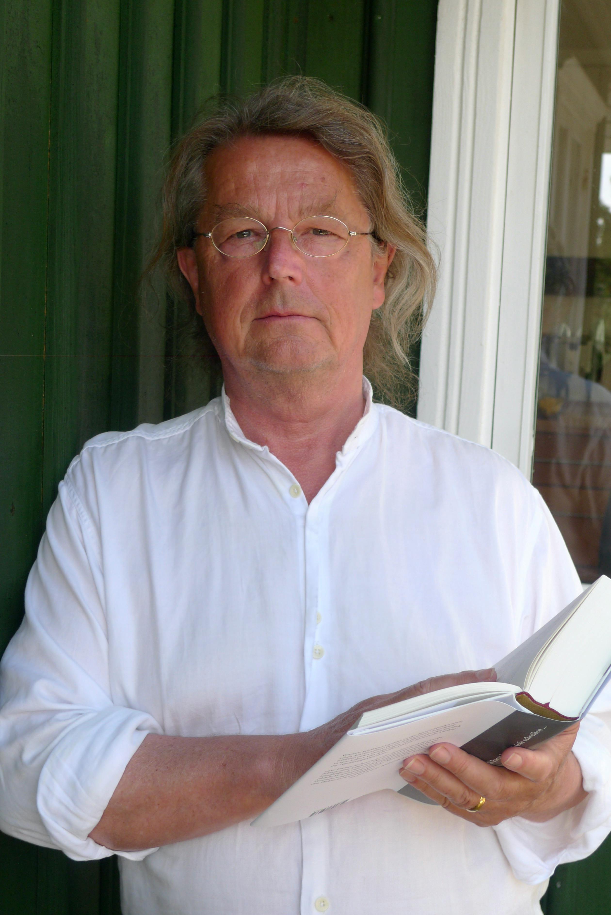 Ein mittelalter Mann mit einem weißen Oberhemd, braunen Haaren und einer Brille hält ein aufgeschlagenes Buch in der Hand.