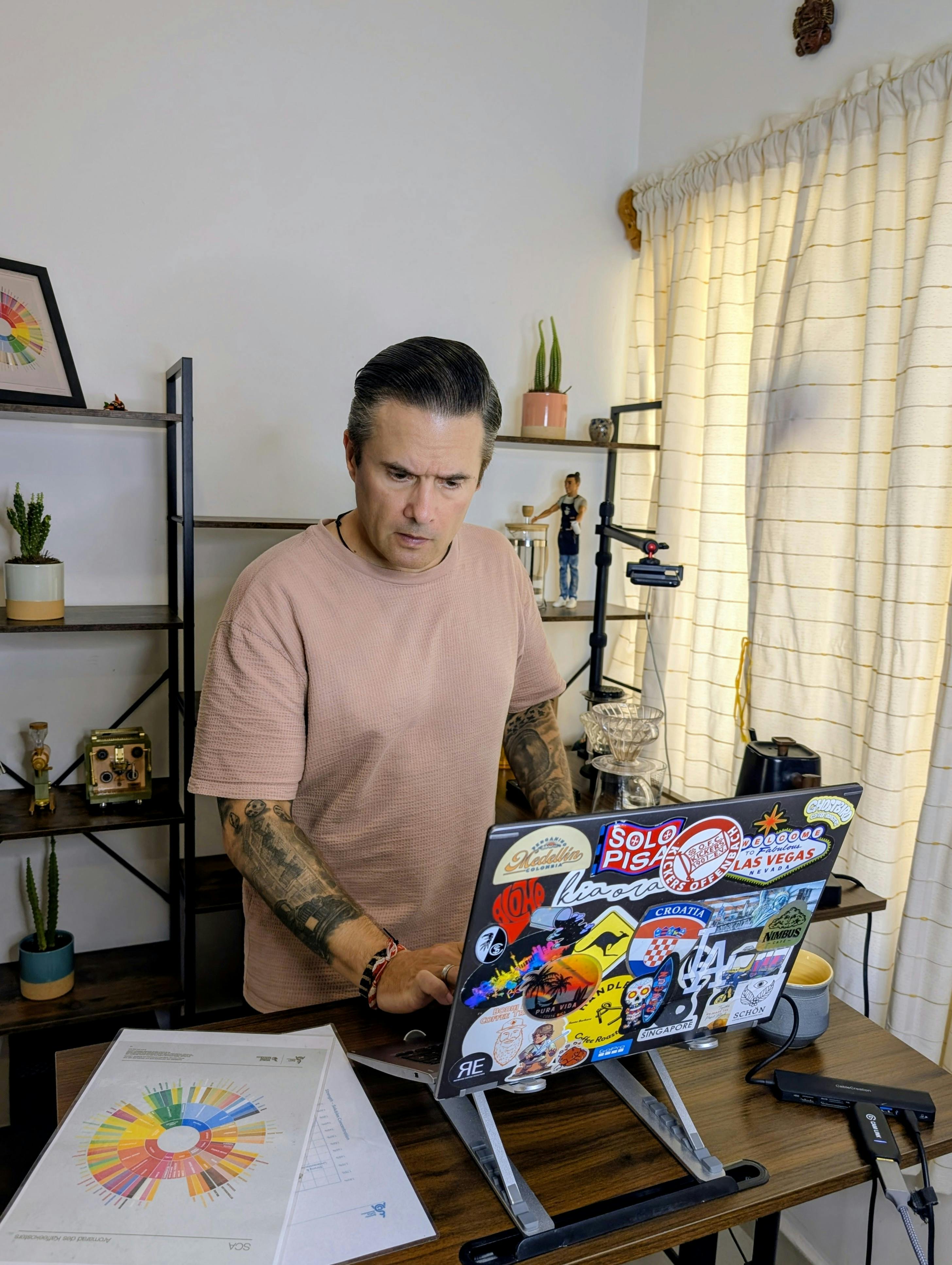 SCA Brewing Professional Daniel (@coffeeteller) konzentriert am Laptop-Arbeitsplatz im Home-Office. Vor ihm auf dem Tisch liegt ein gedrucktes SCA-Aromarad (Flavour Wheel), im Hintergrund Regale mit professionellem Barista-Equipment. Er tippt auf der Tastatur und bereitet ein interaktives Live-Kaffeecoaching via Google Meet für ein Remote-Teamevent vor.
