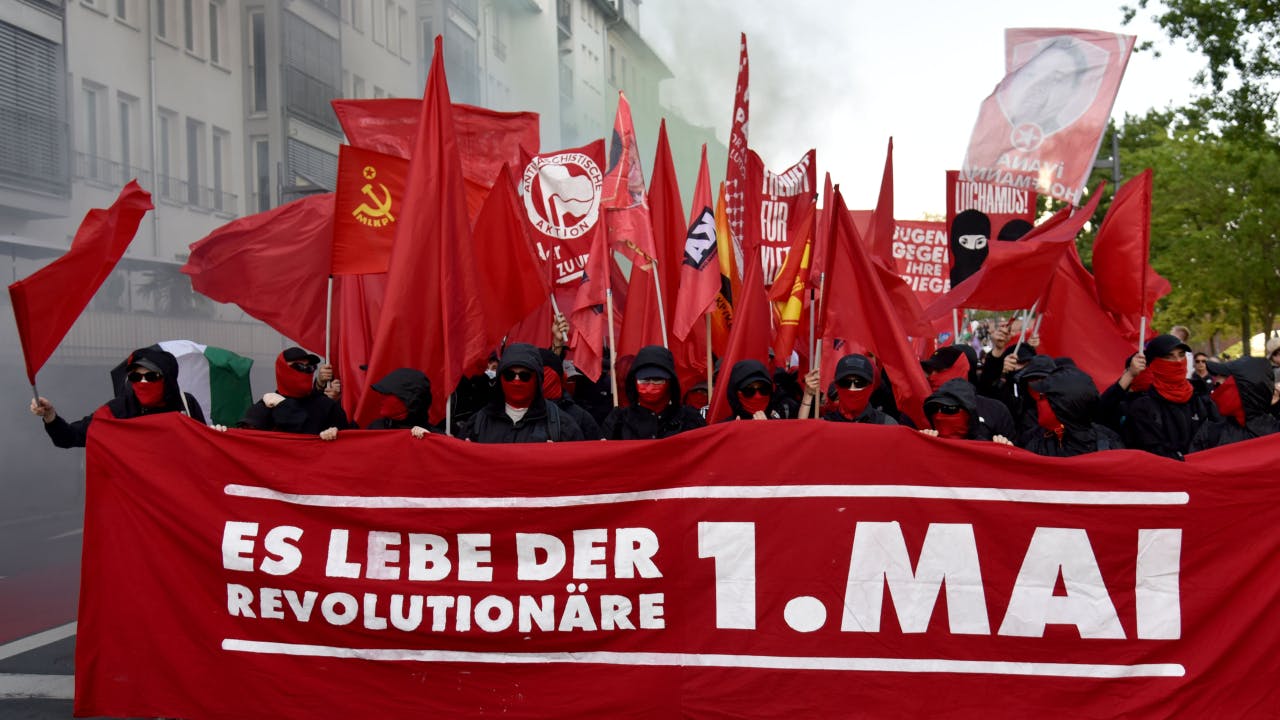 Demozug von vorne fotografiert, rotes Banner "Es lebe der revolutionäre 1. Mai", Menschen in schwarz und rote Flaggen