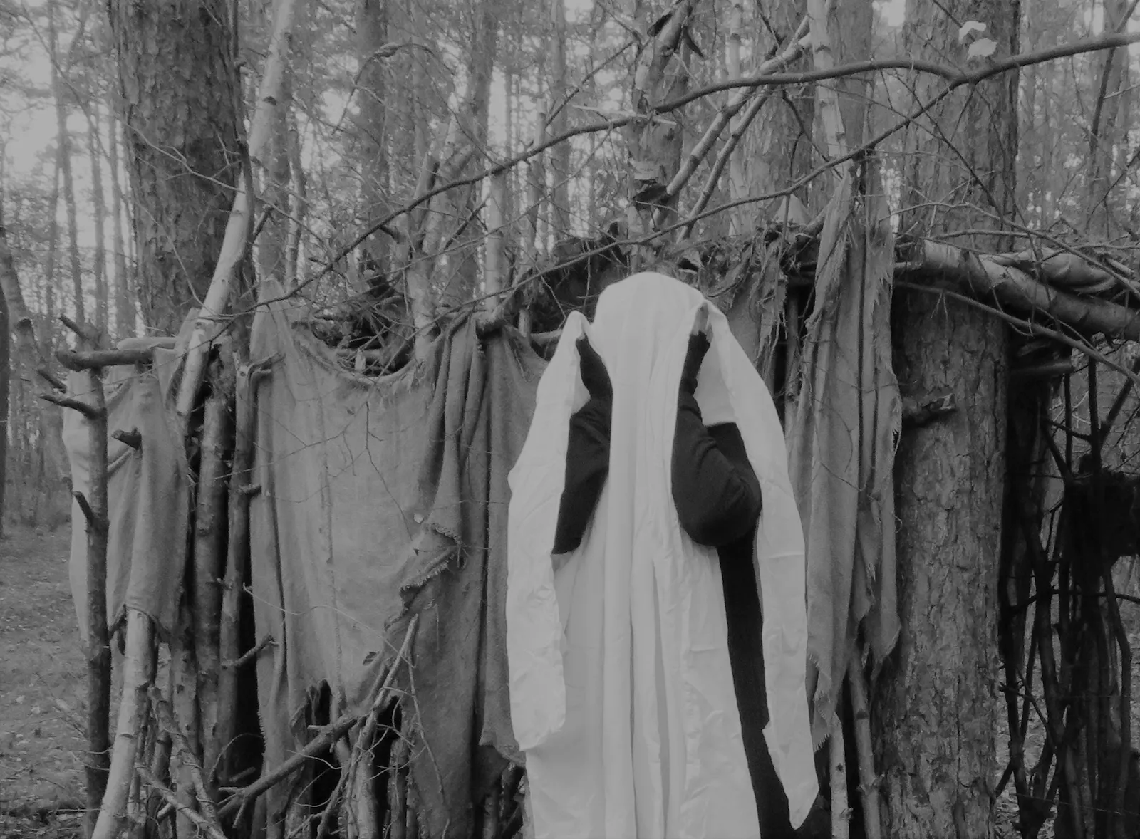 Schwarz-weiße Aufnahme von einer Person unter einem weißen Laken. Sie steht vor einer aus Lumpen und Stöckern gebauten kleinen Hütte im Wald.