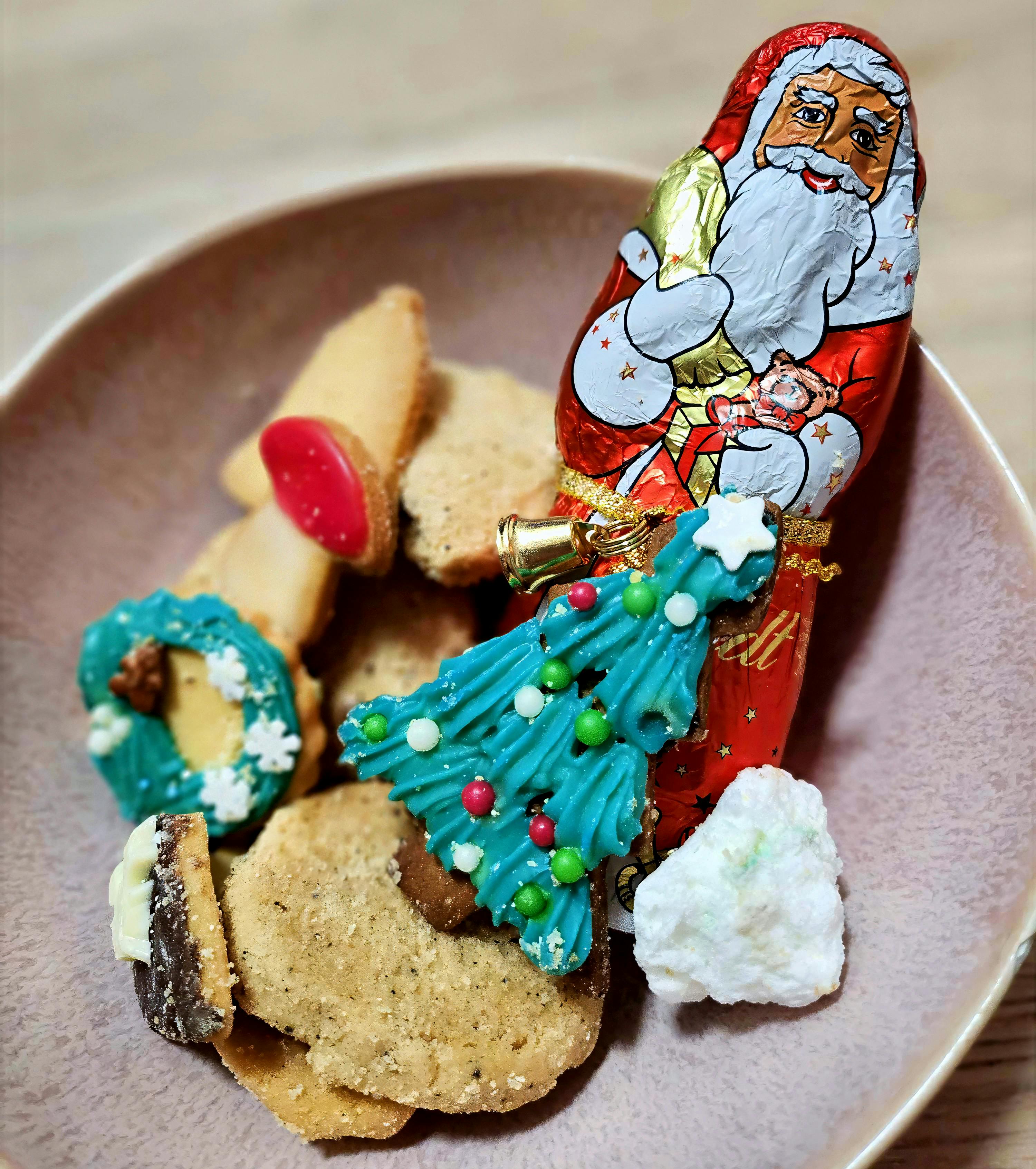 Ein Schokoladen-Nikolaus in roter Folie steht auf einem Teller mit Weihnachtsgebäck. Um ihn herum liegen verschiedene Plätzchen, darunter helle Kekse, ein gefüllter Keks, ein weißes Gebäckstück und ein grün glasierter Tannenbaumkeks mit bunten Zuckerperlen.
