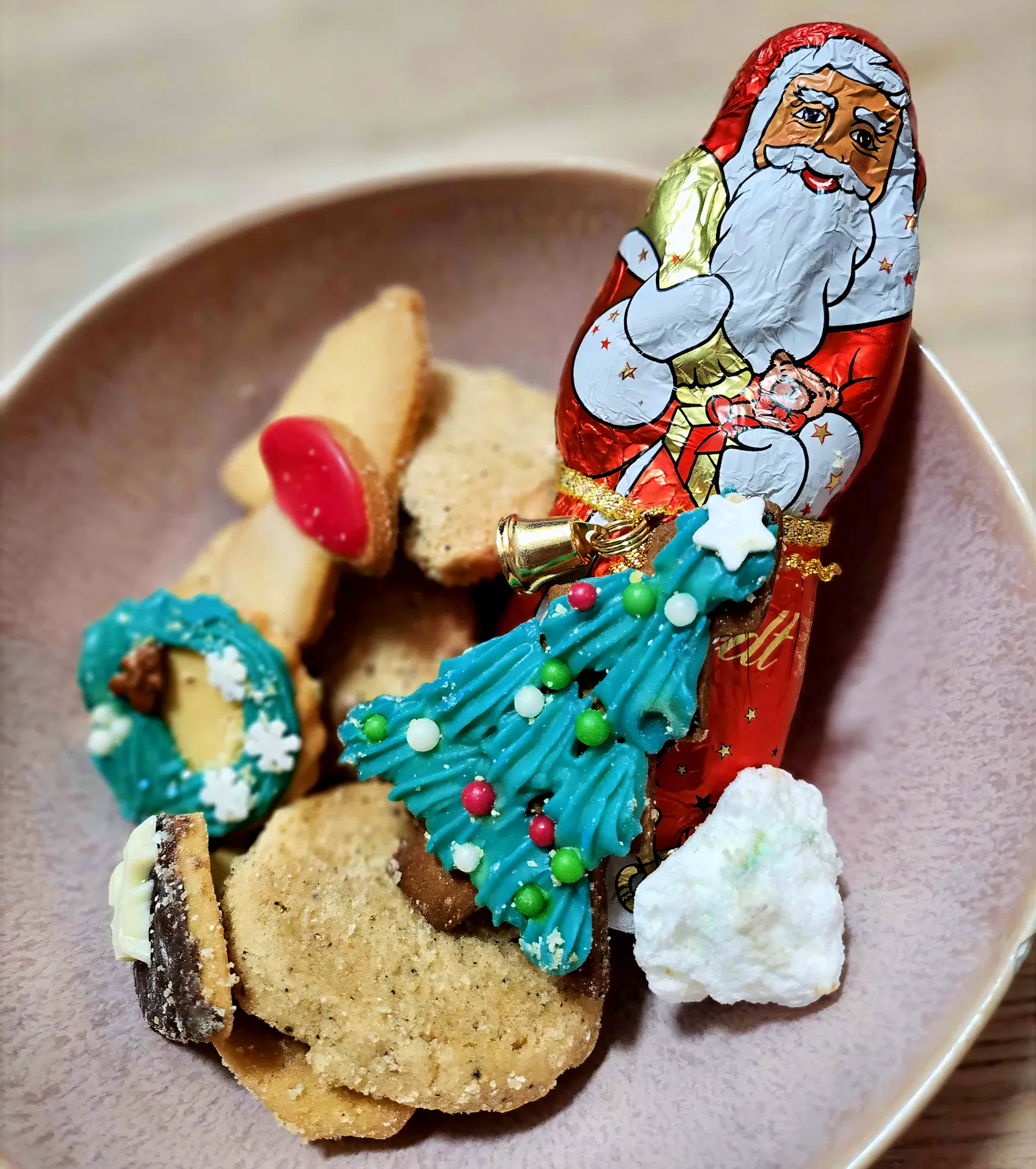 Ein Schokoladen-Nikolaus in roter Folie steht auf einem Teller mit Weihnachtsgebäck. Um ihn herum liegen verschiedene Plätzchen, darunter helle Kekse, ein gefüllter Keks, ein weißes Gebäckstück und ein grün glasierter Tannenbaumkeks mit bunten Zuckerperlen.