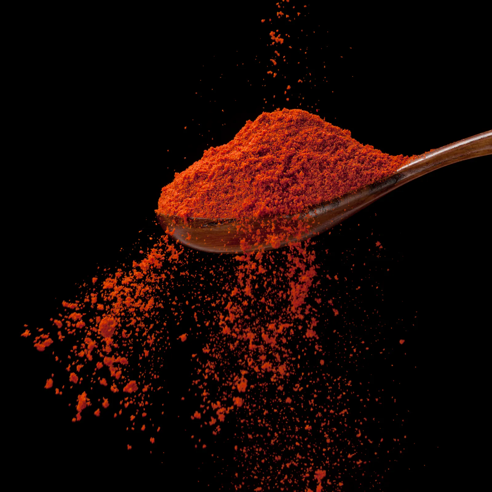 Das Bild zeigt einen Holzlöffel mit leuchtend rotem Paprika-Pulver vor schwarzem Hintergrund, von dem Pulver nach unten rieselt.