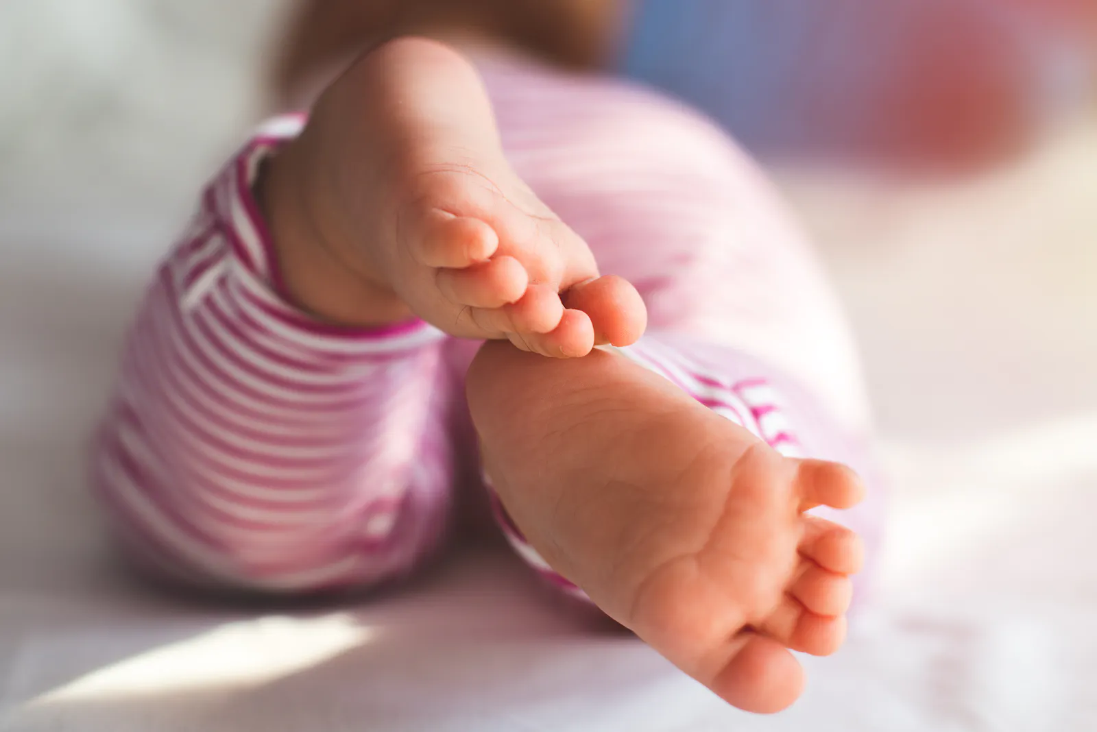 Das Bild zeigt die nackten Füßchen eines Babys in einer rosa-weiß gestreiften Hose aus der Nähe fotografiert.