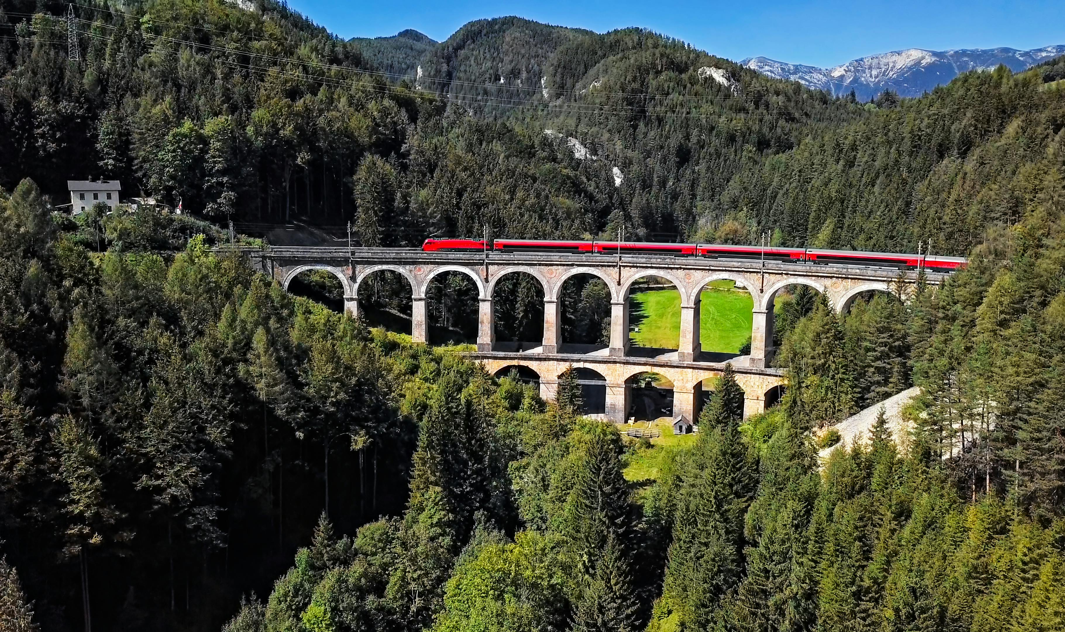 Das Bild zeigt einen roten Zug auf der historischen Semmering-Bahn, der über ein altes Stein-Viadukt fährt, umgeben von dichten Wäldern und Bergen unter blauem Himmel.