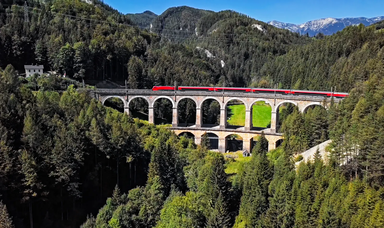 Das Bild zeigt einen roten Zug auf der historischen Semmering-Bahn, der über ein altes Stein-Viadukt fährt, umgeben von dichten Wäldern und Bergen unter blauem Himmel.
