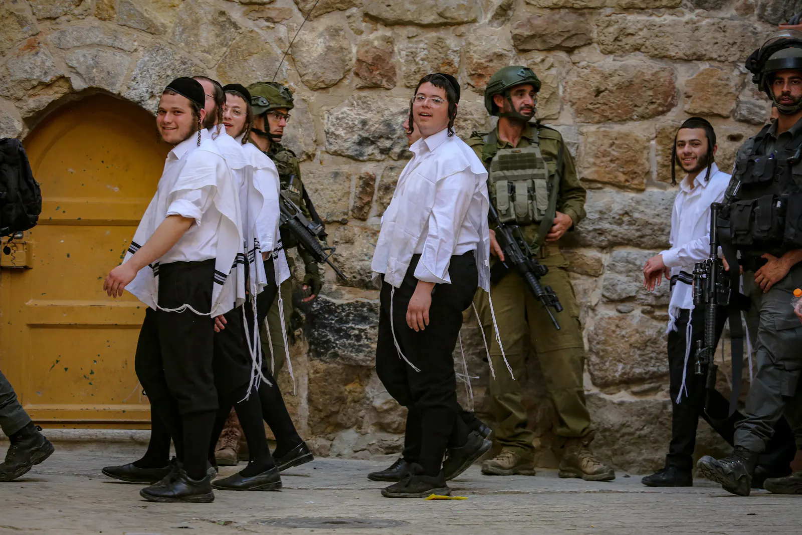 Lächelnde junge Siedler in traditioneller religiöser Kleidung, eskortiert von Soldaten.