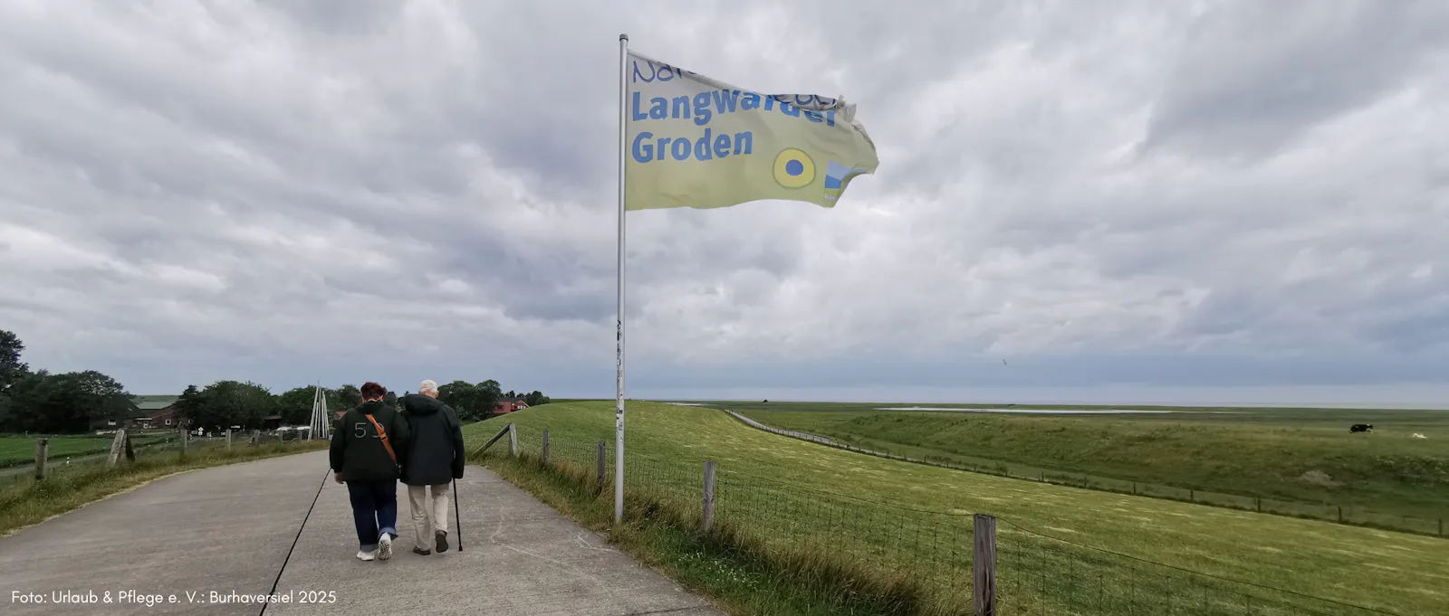 Zwei Personen gehen den Weg vom Langwarder Groden zurück zum Parkplatz. Neben ihnen weht die Fahne des Naturparks im Wind.