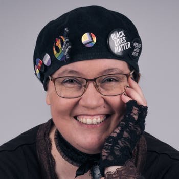 Foto: Amalia trägt eine schwarzen Baskenmütze mit mehreren Buttons und eine Brille. They lächelt den Betrachter an und hat eine Hand mit einem Rüschenhandschuh an their Wange gelegt.