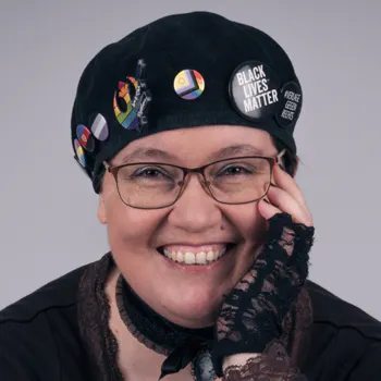 Foto: Amalia trägt eine schwarzen Baskenmütze mit mehreren Buttons und eine Brille. They lächelt den Betrachter an und hat eine Hand mit einem Rüschenhandschuh an their Wange gelegt.