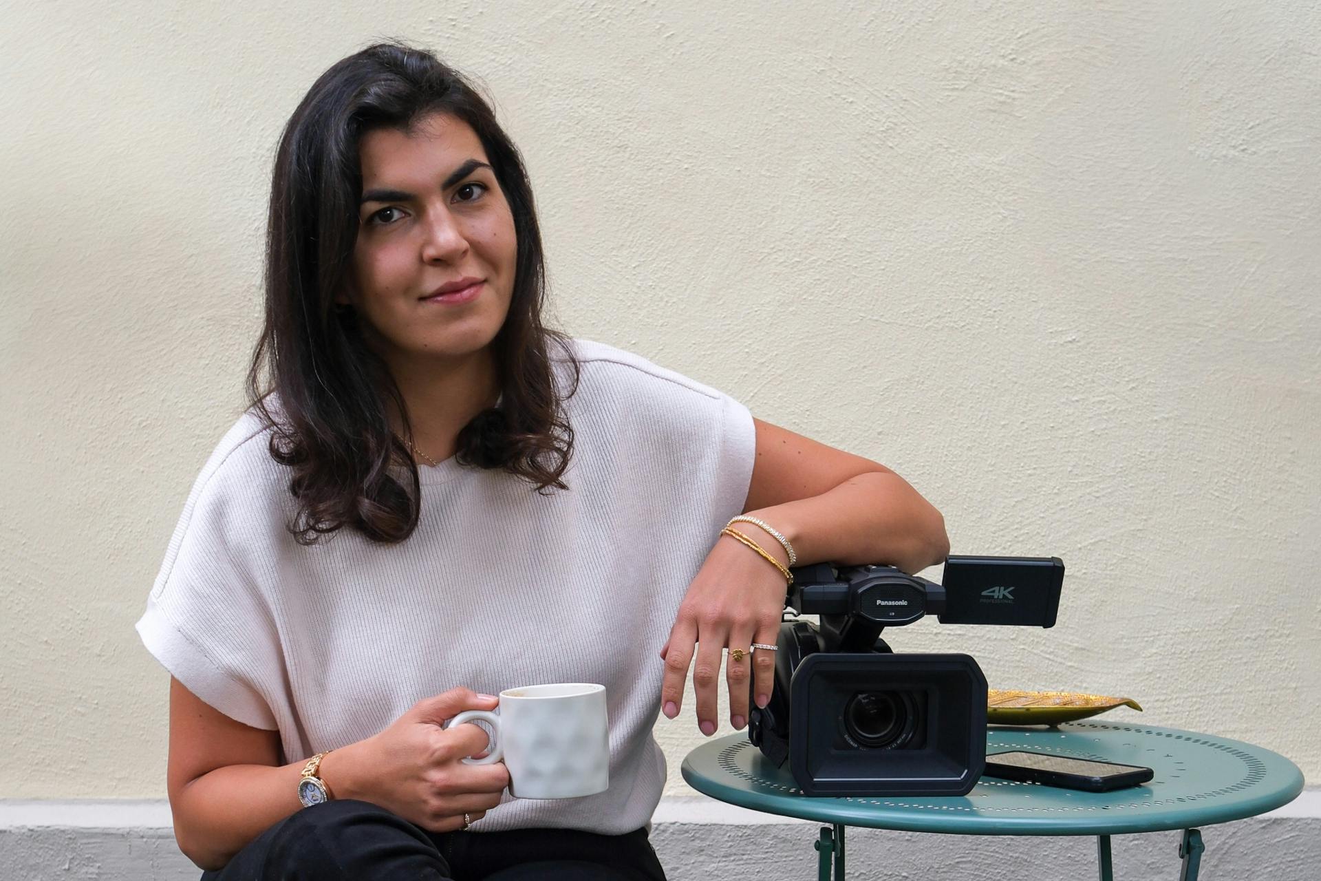 Ayla Yildiz hält eine Tasse in der einen Hand und der andere Arm lehnt auf einer Kamera, die auf einem Tisch liegt. Sie trägt ein helles, kurzes Oberteil.