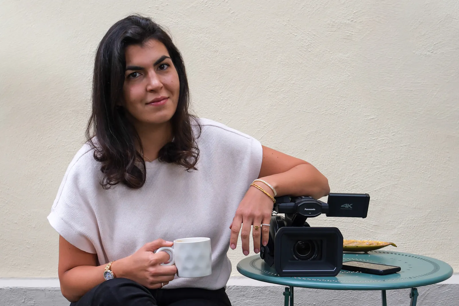 Ayla Yildiz hält eine Tasse in der einen Hand und der andere Arm lehnt auf einer Kamera, die auf einem Tisch liegt. Sie trägt ein helles, kurzes Oberteil.