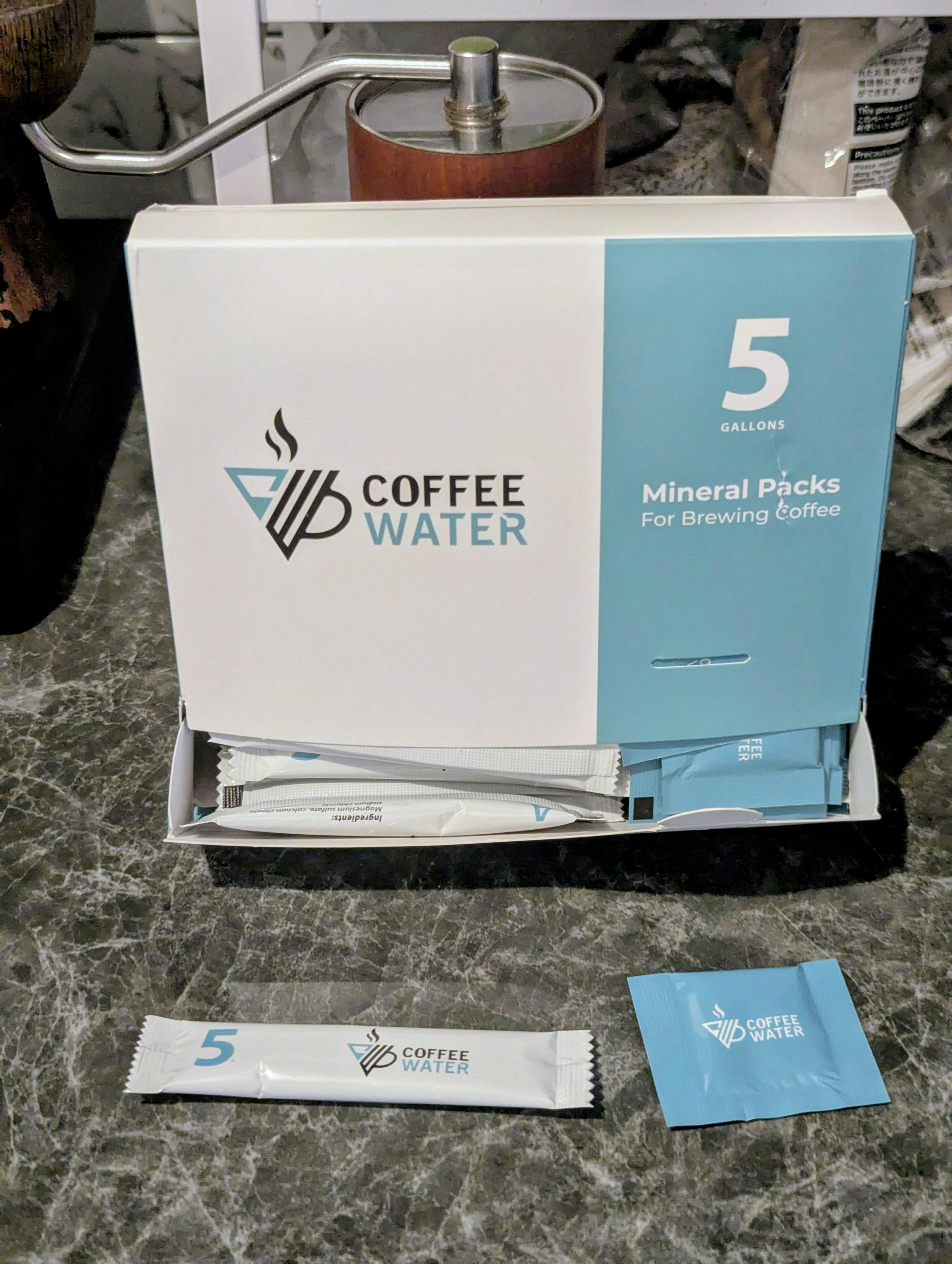 Eine geöffnete Schachtel ‚Coffee WATER Mineral Packs‘ mit zwei verschiedenen Mineraliensäckchen (Stift- und Quadratformat) davor. Dieses Bild zeigt die Mineralien, die nötig sind, um Kalk und ungefilterte Mineralien im Kaffeewasser zu ersetzen.
