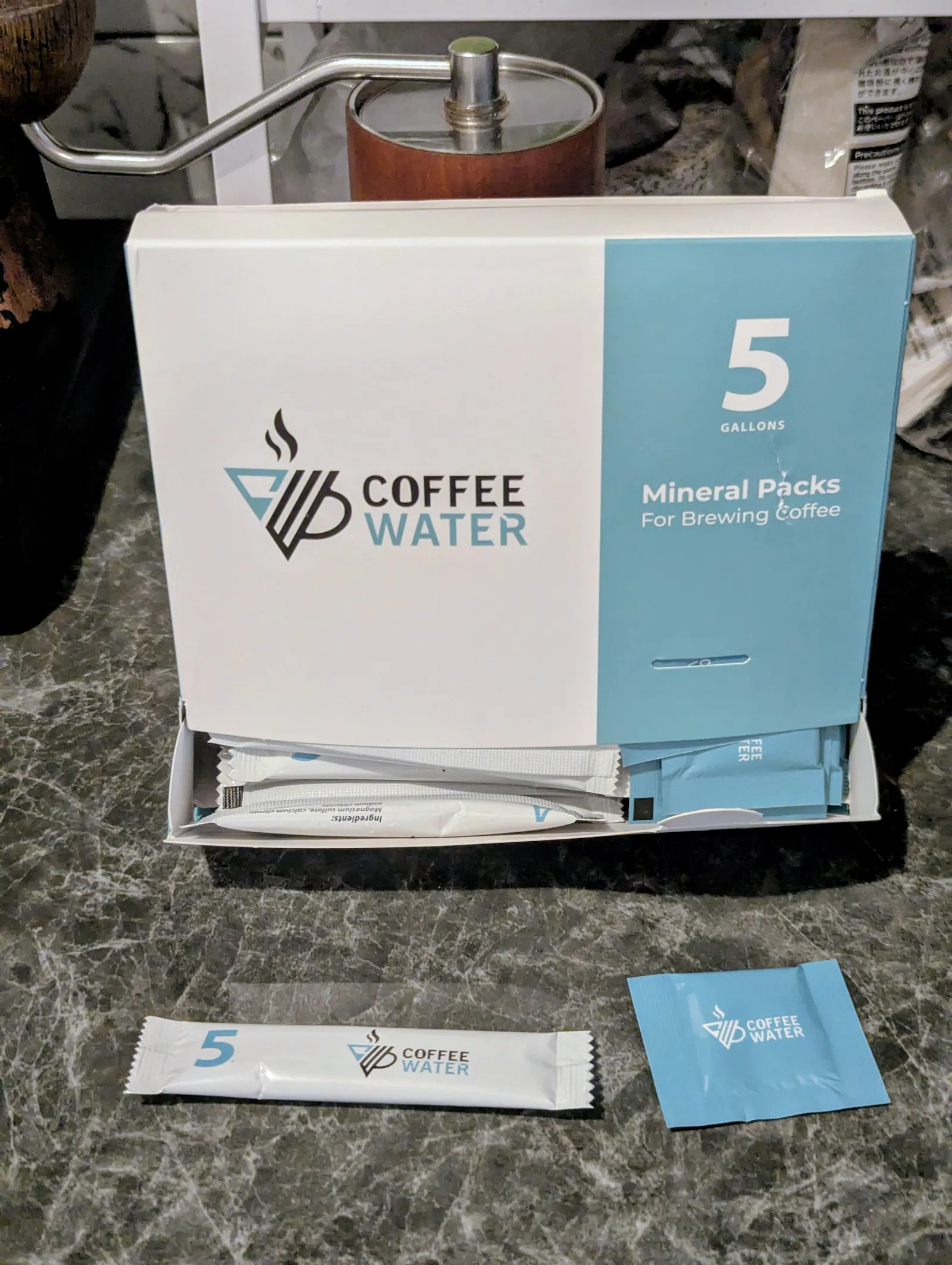 Eine geöffnete Schachtel ‚Coffee WATER Mineral Packs‘ mit zwei verschiedenen Mineraliensäckchen (Stift- und Quadratformat) davor. Dieses Bild zeigt die Mineralien, die nötig sind, um Kalk und ungefilterte Mineralien im Kaffeewasser zu ersetzen.