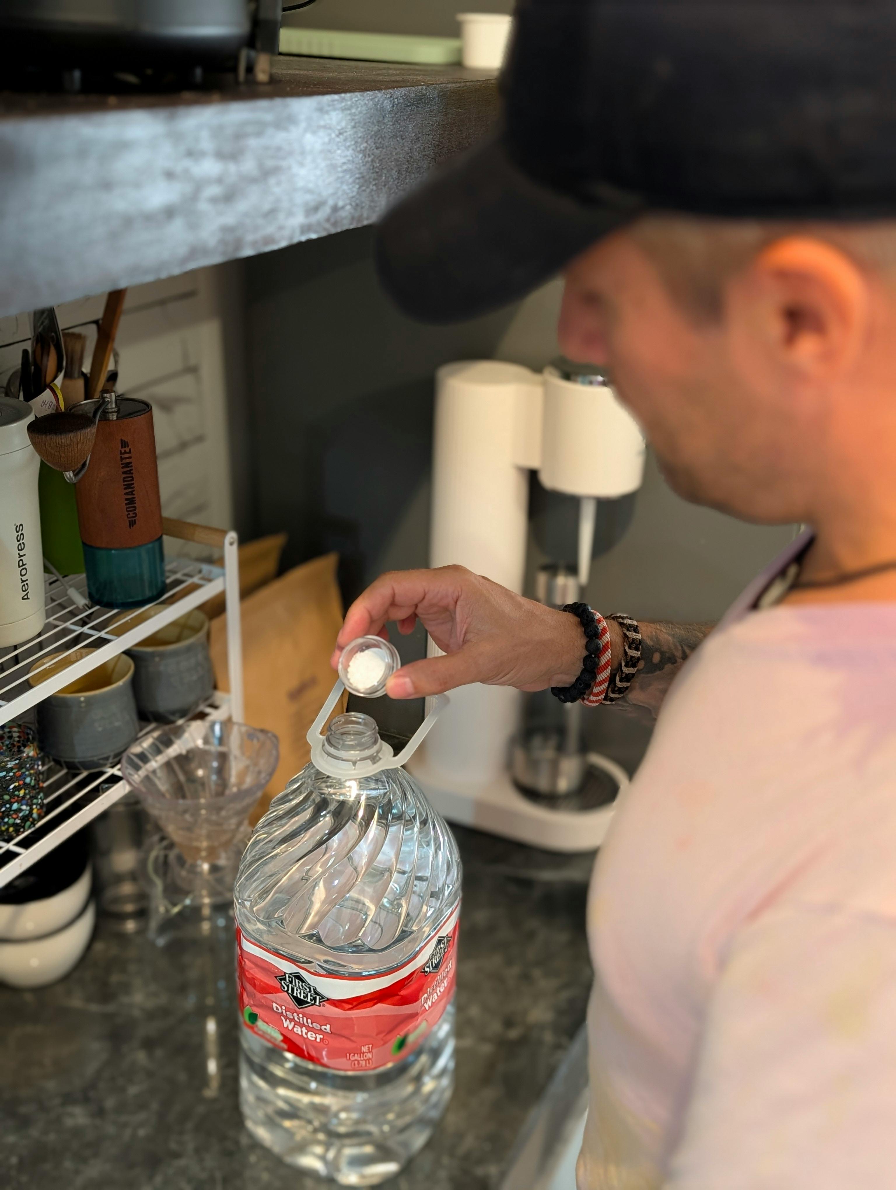 Nahaufnahme von Daniel Frank (Coffeeteller) in Mütze und T-Shirt, der Mineralienpulver aus einem kleinen Behälter in eine große 1-Gallonen-Wasserflasche fügt. Dies verdeutlicht den persönlichen, handwerklichen Prozess der Wasseroptimierung für Specialty Coffee.