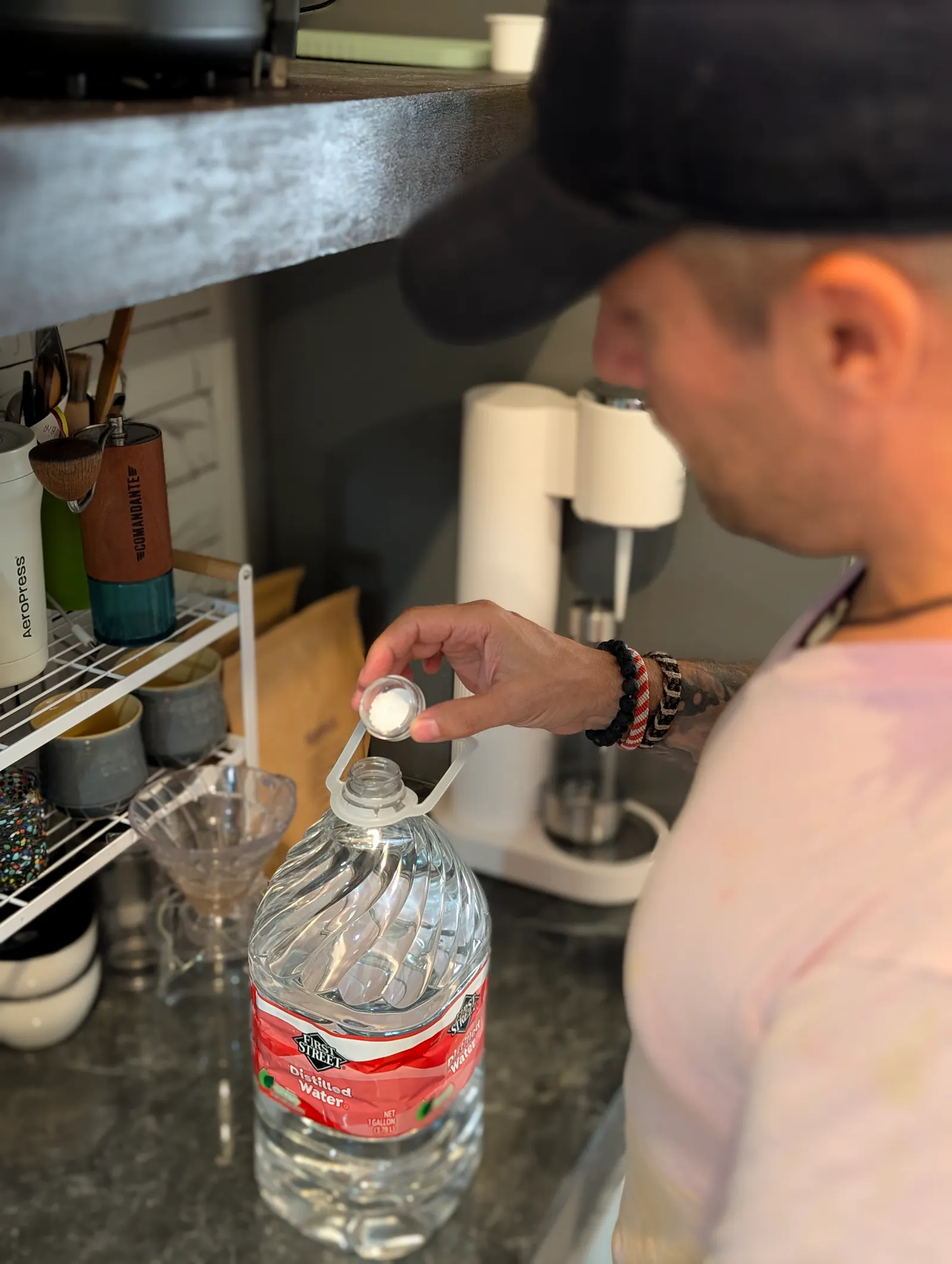 Nahaufnahme von Daniel Frank (Coffeeteller) in Mütze und T-Shirt, der Mineralienpulver aus einem kleinen Behälter in eine große 1-Gallonen-Wasserflasche fügt. Dies verdeutlicht den persönlichen, handwerklichen Prozess der Wasseroptimierung für Specialty Coffee.