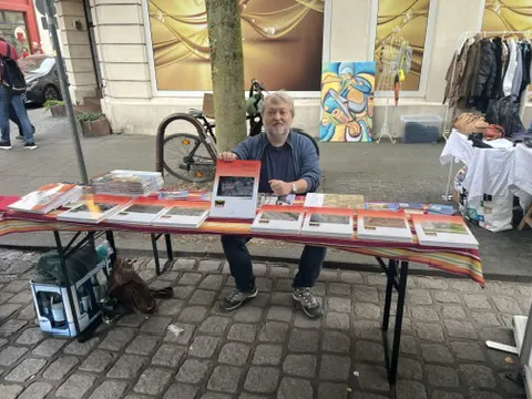 Ein Mensch sitzt hinter einem Büchertisch und hält ein Buch hoch.