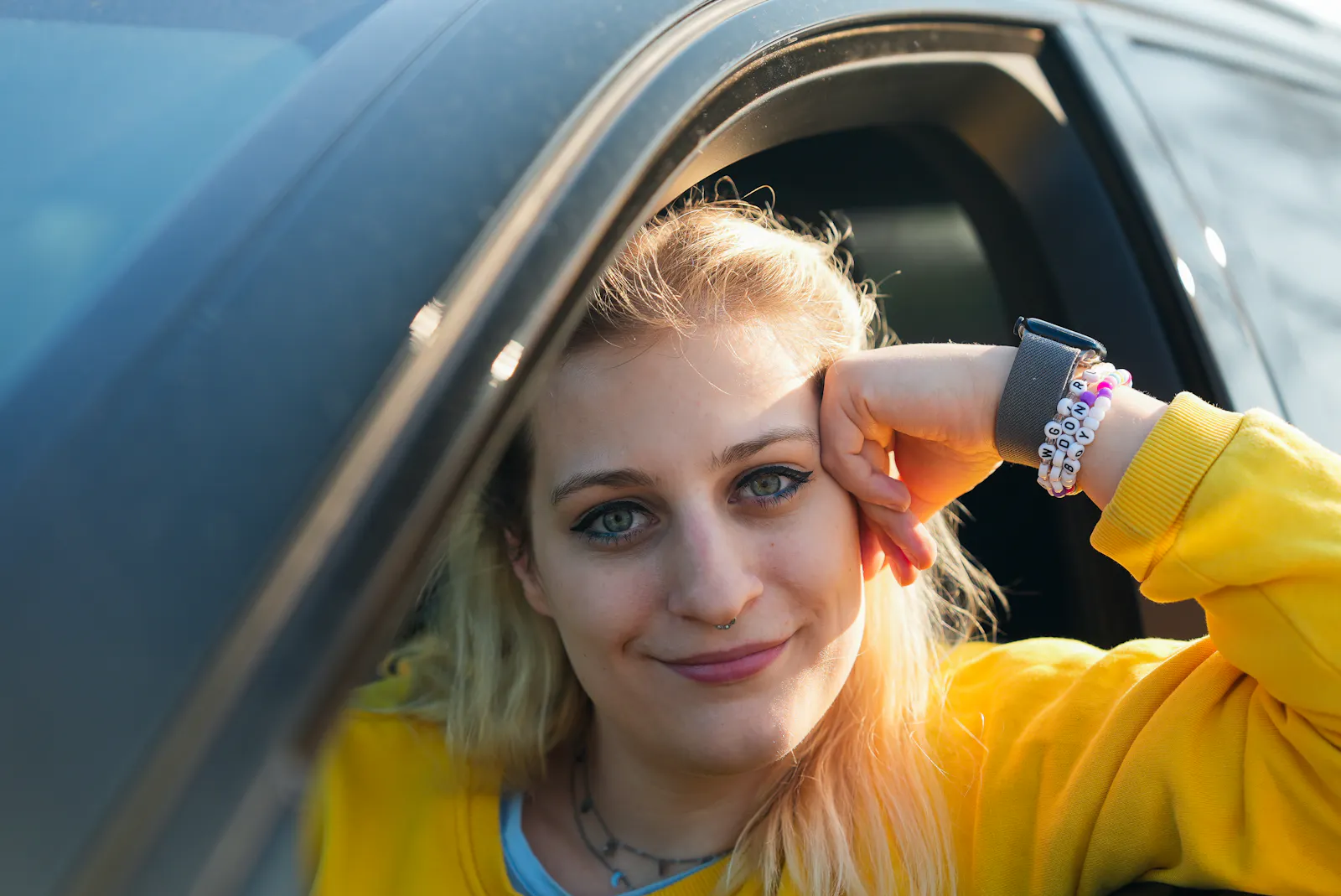 Eine junge Frau in einem gelben Pullover schaut aus dem Fenster eines schwarzen Wagens, Fahrerseite. Am Arm trägt sie eine Apple Watch sowie zwei Freundschaftsbändchen im Taylor Swift Style.