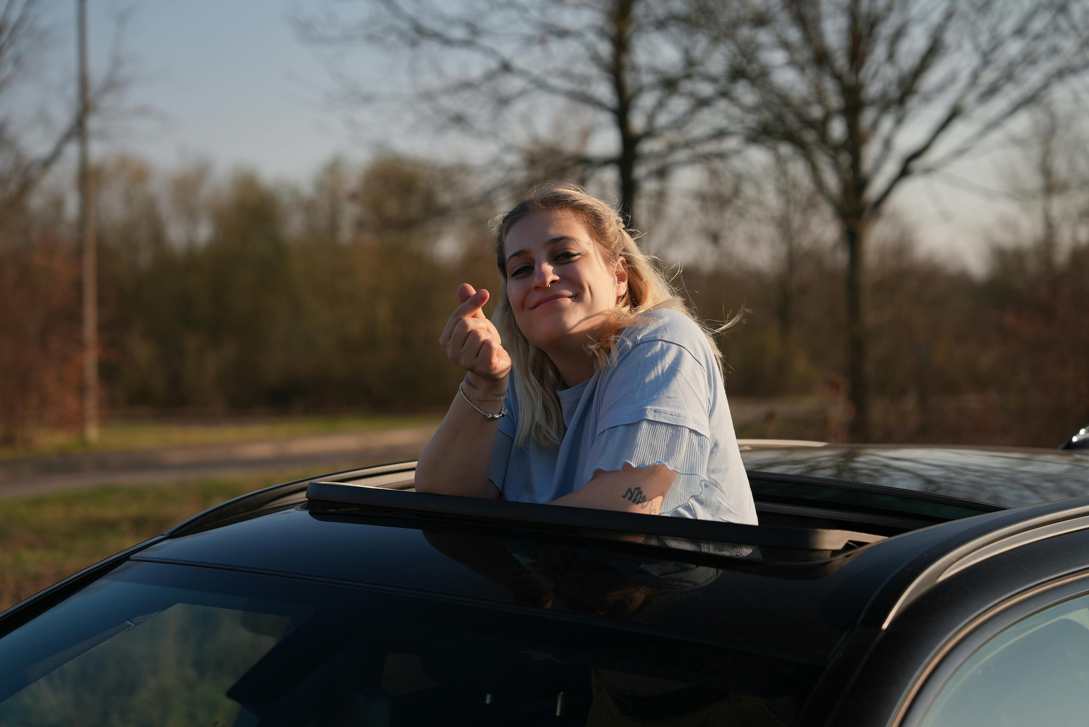 Eine junge blonde Frau, die aus dem Dachfenster eines schwarzen Fahrzeuges schaut, sich entspannt an den Rand lehnt in einem blauen T-Shirt, in die Kamera lächelt und dabei mit ihrer rechten Hand ein kleines K-Pop Herz formt.
