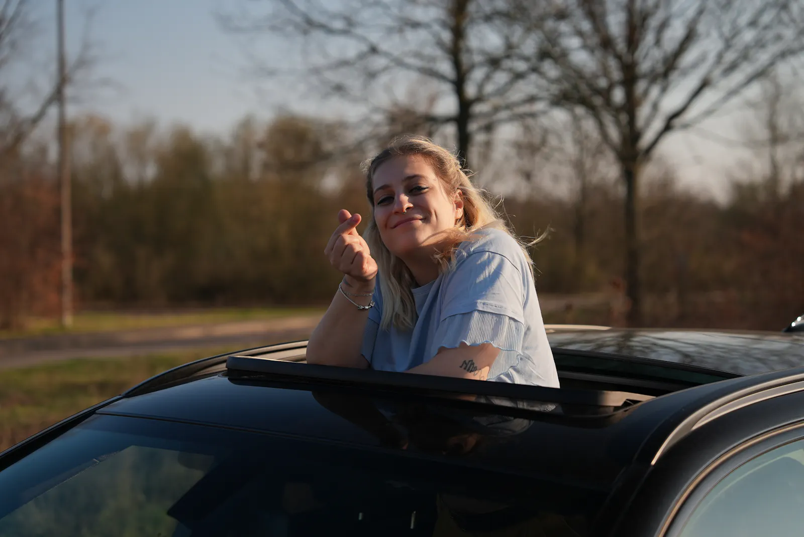 Eine junge blonde Frau, die aus dem Dachfenster eines schwarzen Fahrzeuges schaut, sich entspannt an den Rand lehnt in einem blauen T-Shirt, in die Kamera lächelt und dabei mit ihrer rechten Hand ein kleines K-Pop Herz formt.