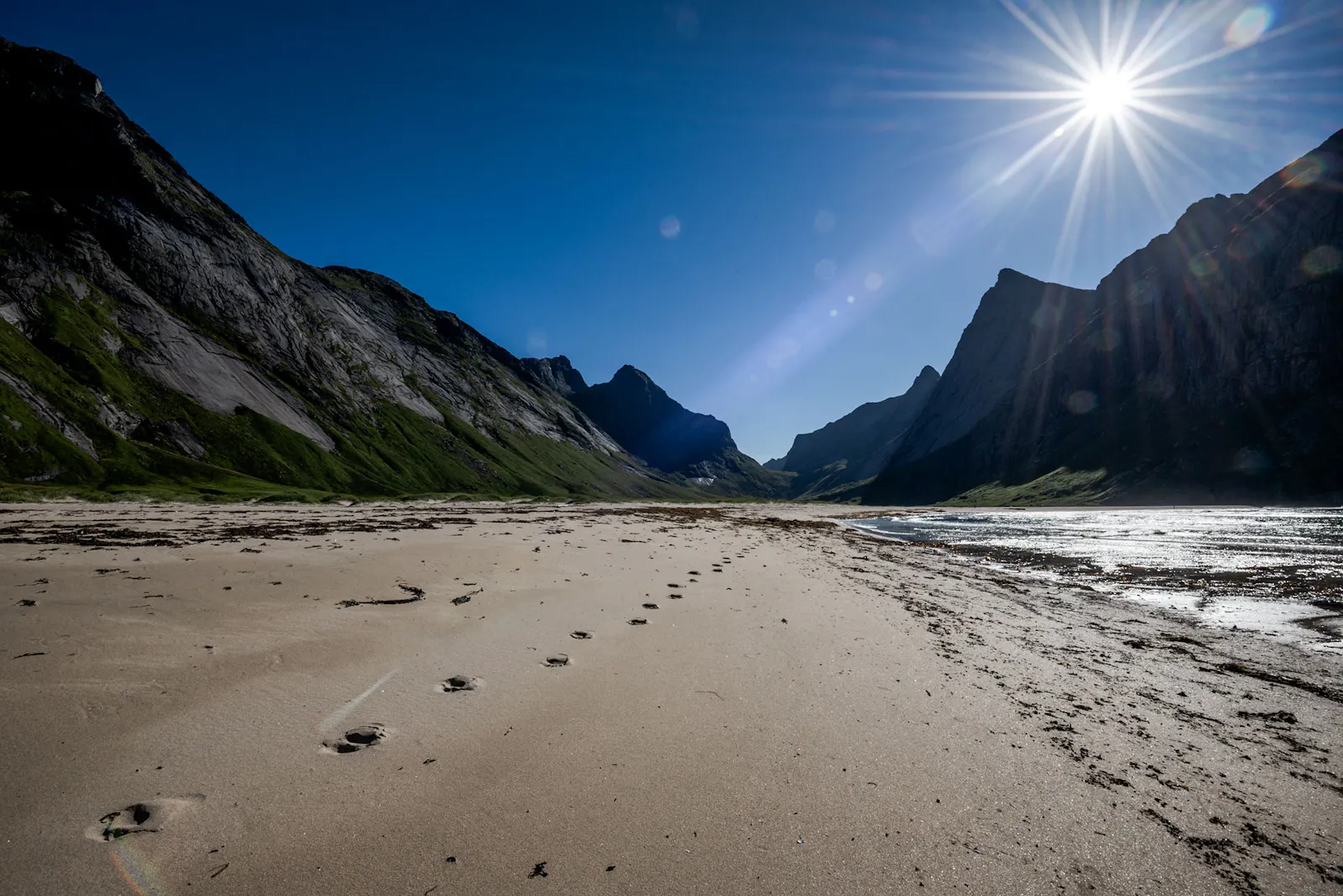 La plage au premier plan, avec une série de traces de pieds nus qui part au loin. En arrière-plan, les chaînes de montagne de la vallée, qui se rejoignent en un col tout au fond.