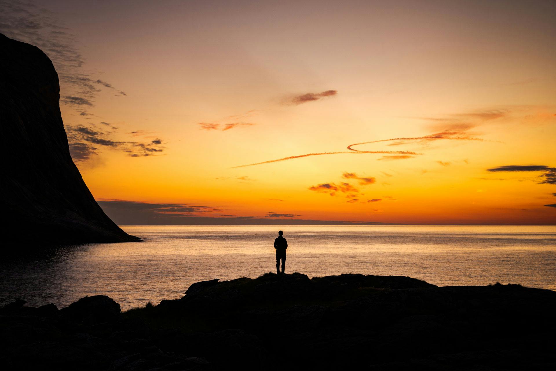 Une silhouette humaine en contre-jour d'un magnifique coucher de soleil, debout sur un promontoire au-dessus de la mer qui semble infinie. À sa gauche, une falaise qui tombe dans l'eau.