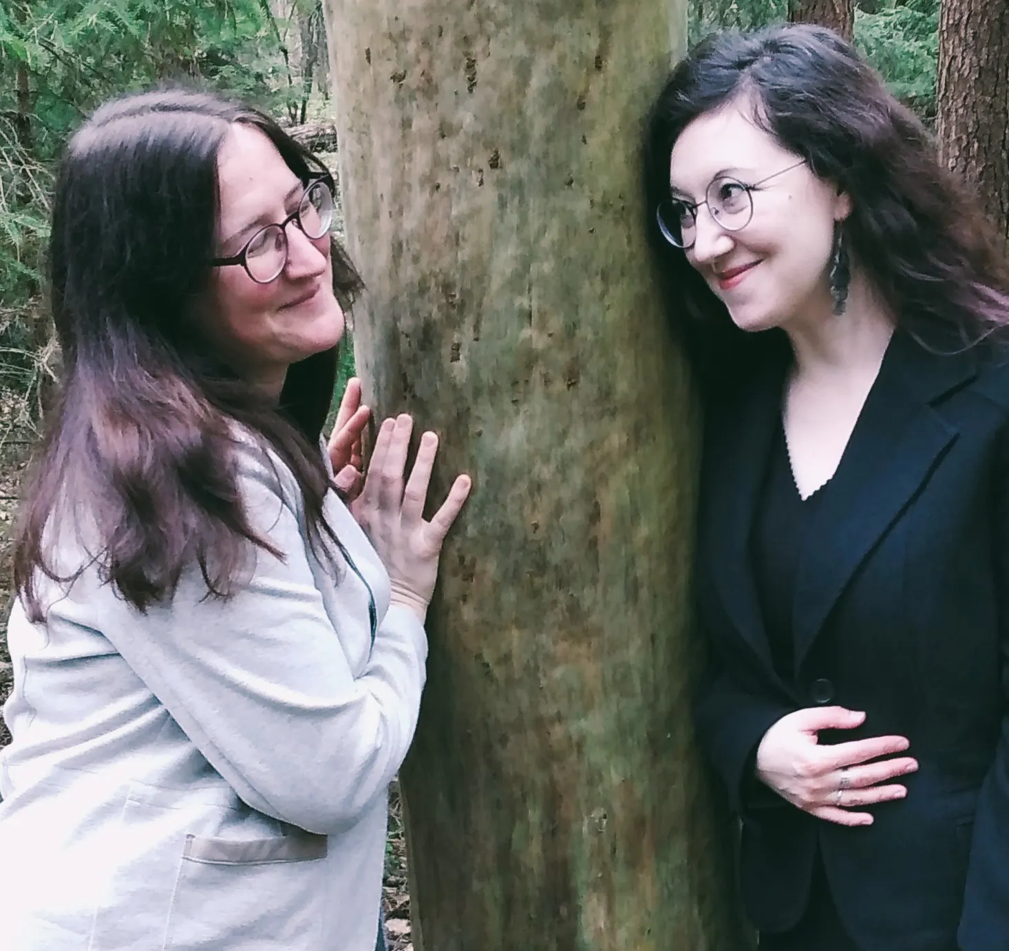 Zwei Frauen stehen an einen Baum gelehnt im Wald. Eine trägt helle, die andere dunkle Kleidung. Sie blicken einander freundlich an und lächeln.