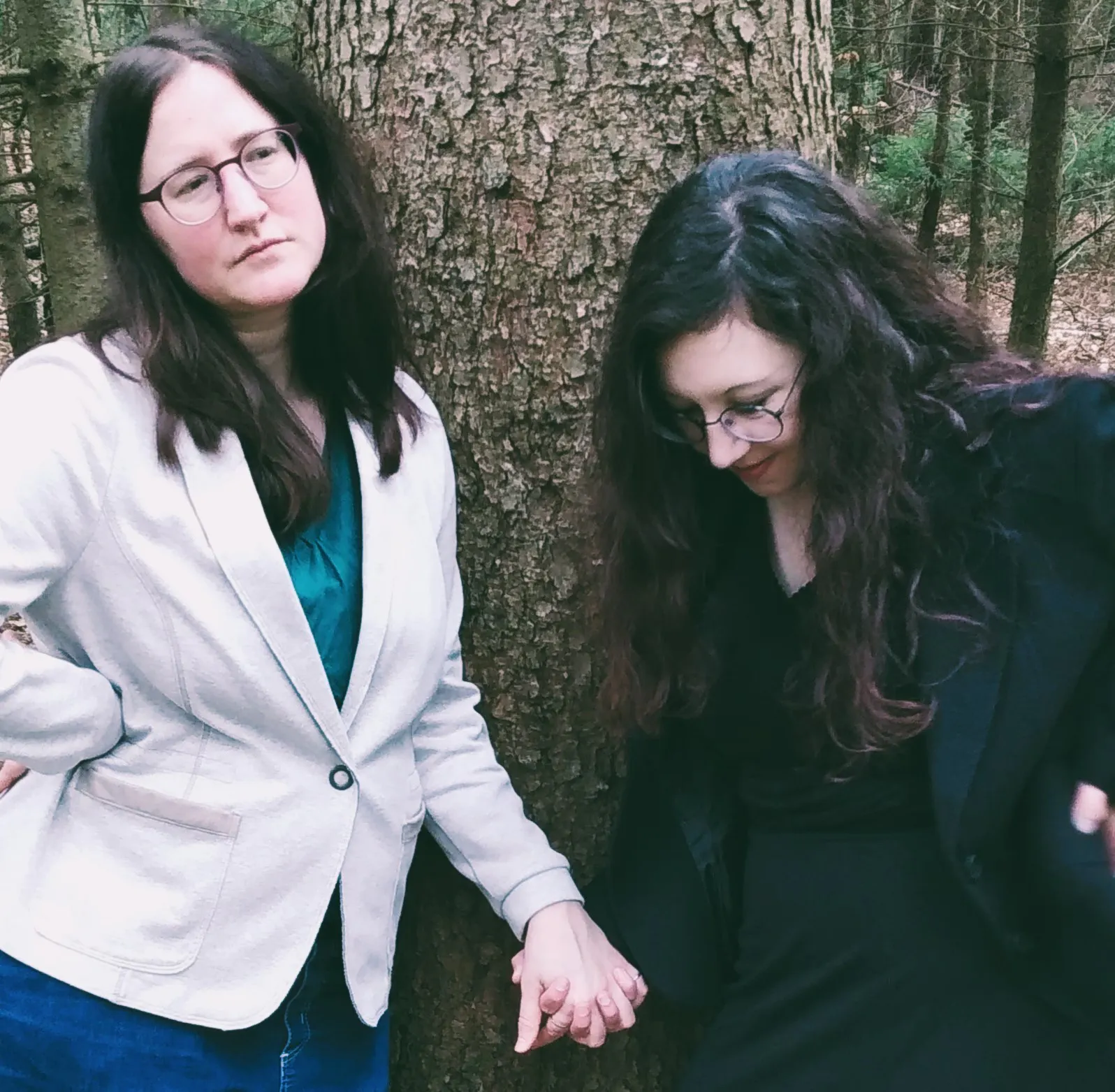 Zwei Frauen stehen an einen Baum gelehnt im Wald. die Frauen halten sich an den Händen. Eine von ihnen ist in hellen Tönen, die andere in dunklen gekleidet. Eine sieht geradewegs in die Ferne, die andere nach unten.