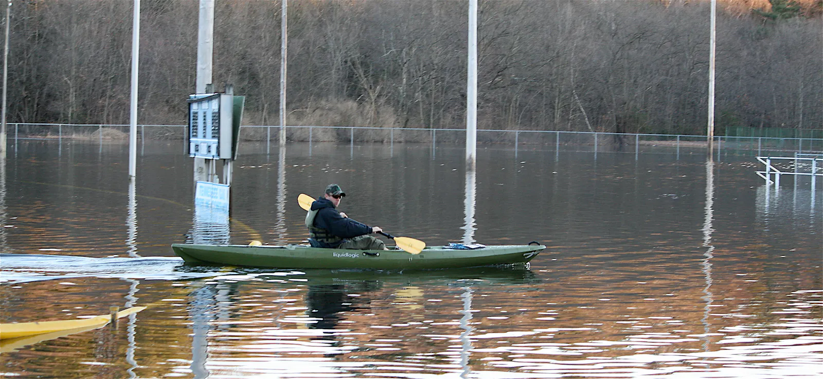 A flooded baseball field in March of 2010 with the water levels up beneath the scoreboard. A man in a black sweatshirt and green hat kayaks over the flooded field in a green kayak with yellow paddles. The bleachers on the right side of the field have water up to the second to last seats.