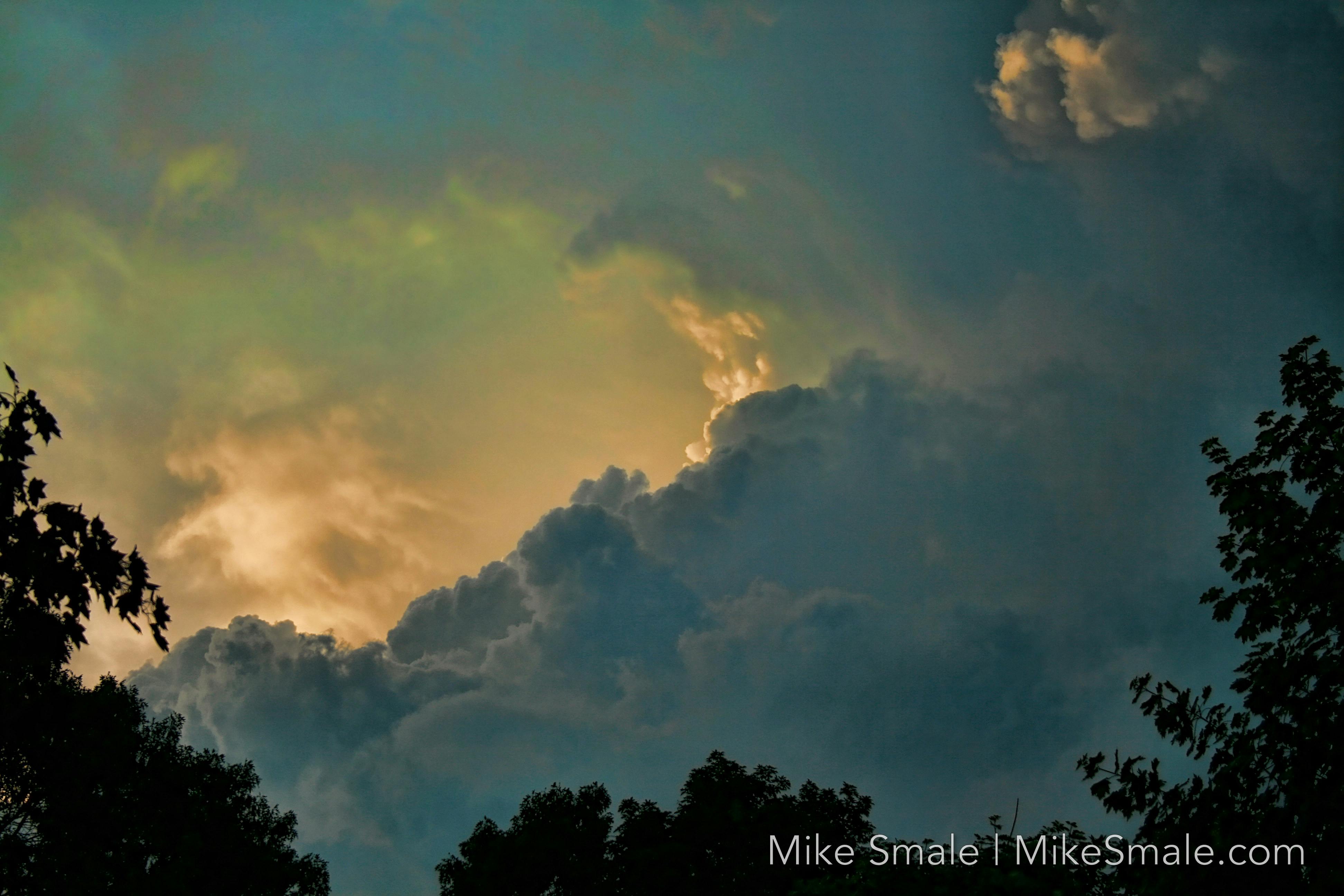 Storm clouds gather as the sun sets, casting a golden glow. Dark silhouettes of trees frame the sky, enhancing the scene's drama.