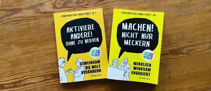 Zwei Arbeitshefte für eine bessere Welt liegen auf einer Holztischplatte.