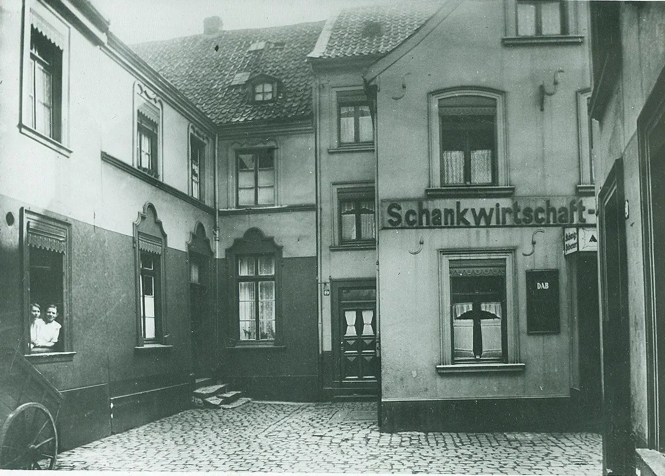 Schwarz-Weiß Foto einer verwinkelter Straße. Zu sehen ist eine "Schankwirtschaft" und aus dem Fenster schauen zwei junge Frauen.
Duisburg-Ruhrort 1920-30er Jahre.