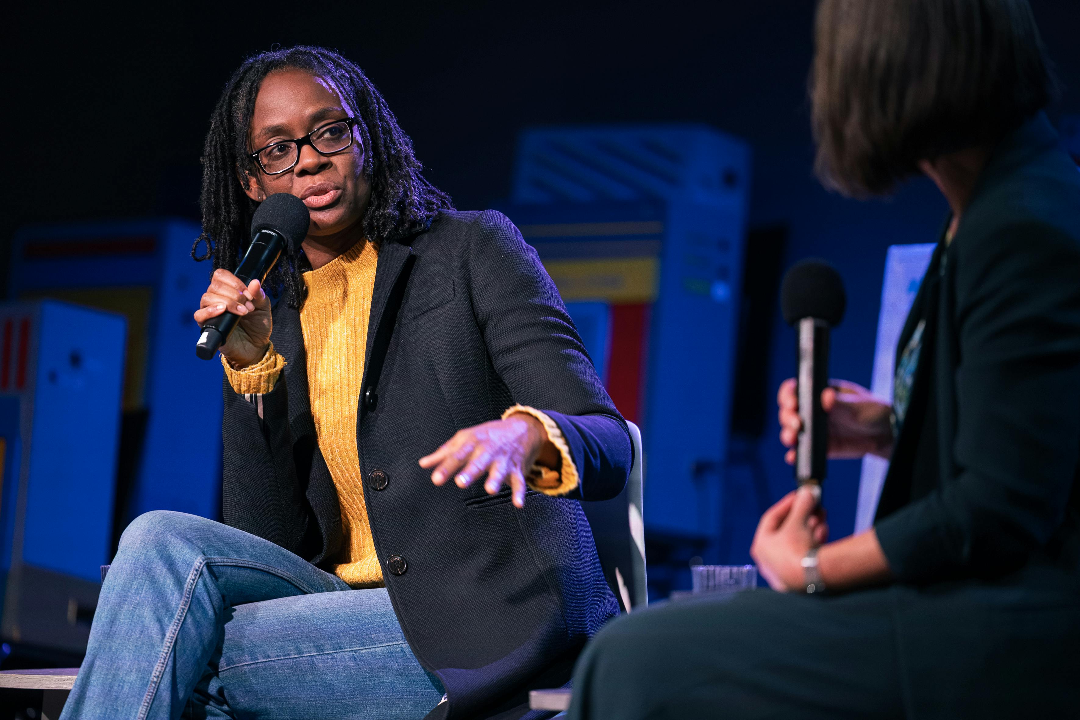 Ein Foto von Sharon Otoo, sie trägt einen gelben Pullover, eine blaue Jeans und eine schwarze Jacke. Sie sitzt auf der Bühne und hält eine Mikrofon in der Hand.