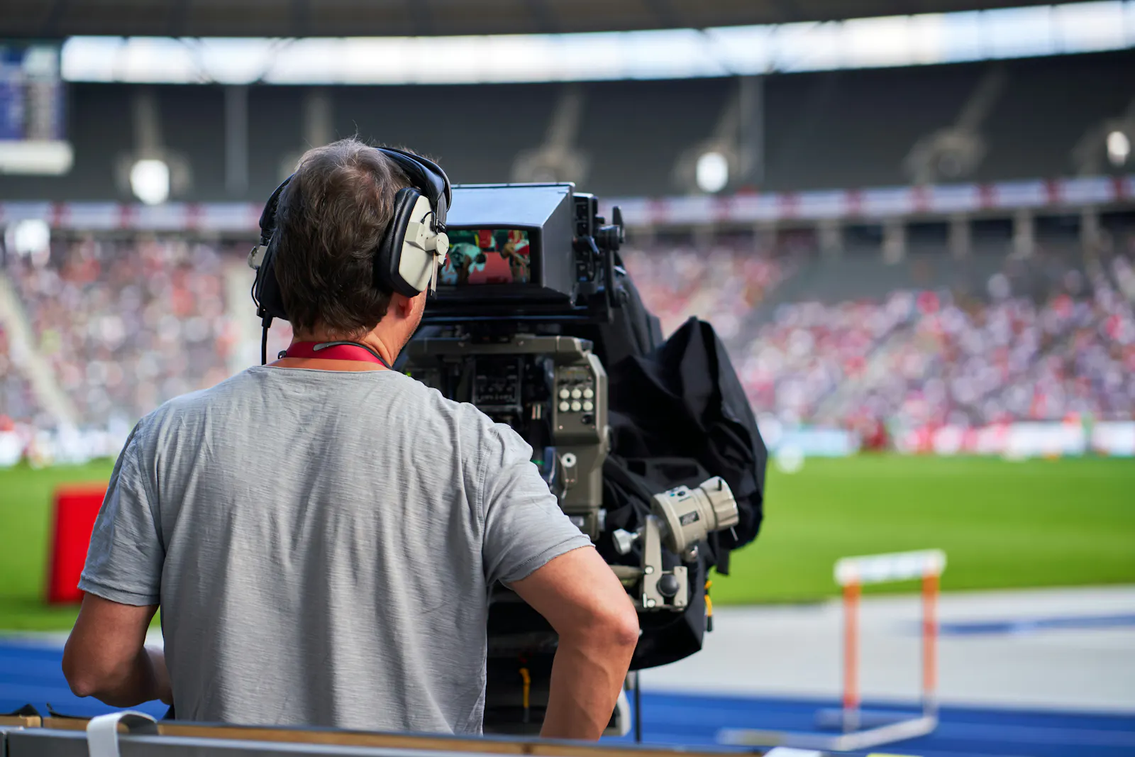 Mann mit Kopfhörern vor einer großen TV-Kamera in einem Stadion