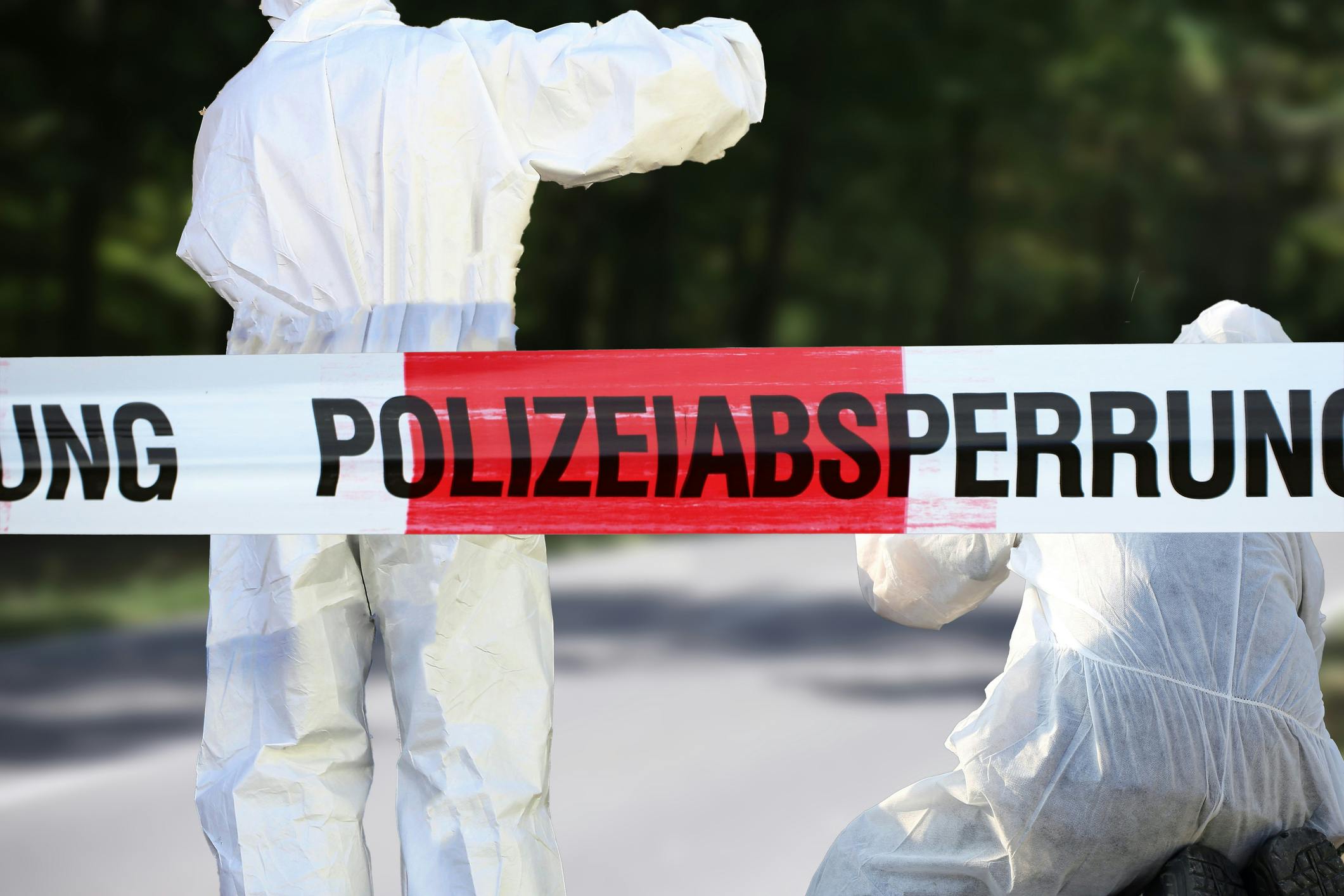 Das Bild zeigt zwei Personen in weißen Schutzanzügen hinter einem rot-weißen Absperrband mit der Aufschrift „Polizeiabsperrung".