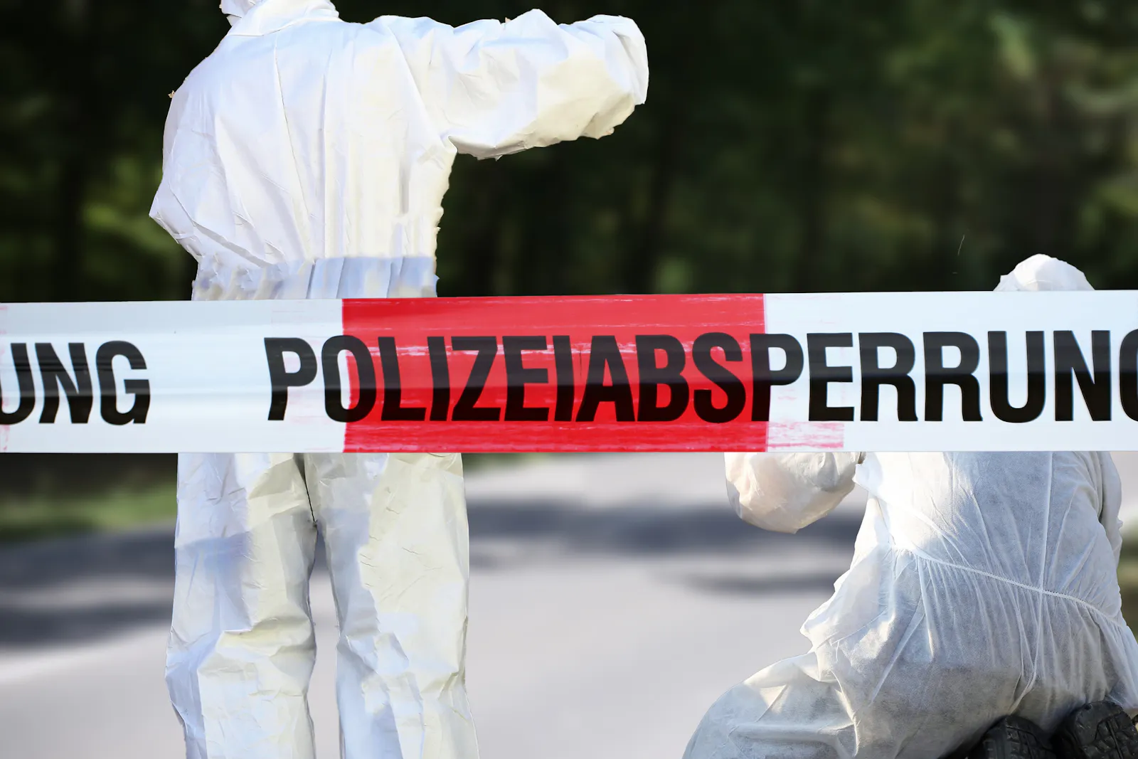 Das Bild zeigt zwei Personen in weißen Schutzanzügen hinter einem rot-weißen Absperrband mit der Aufschrift „Polizeiabsperrung".