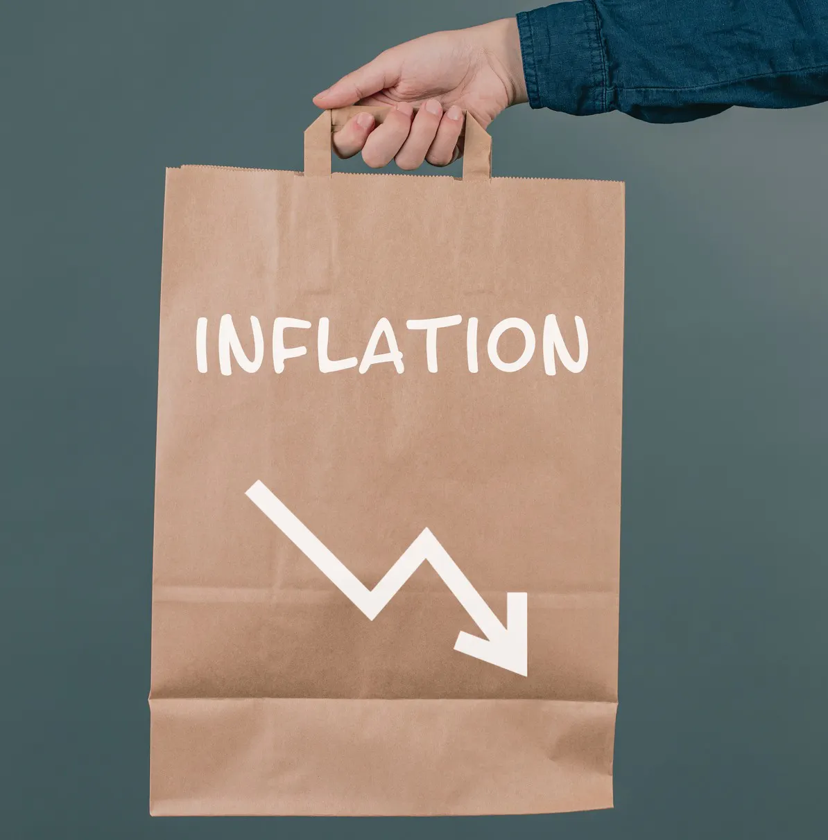 Das Bild zeigt eine Hand, die eine braune Papiereinkaufstasche hält. Auf der Tasche steht das Wort „Inflation“ und ein Pfeil zeigt nach unten.