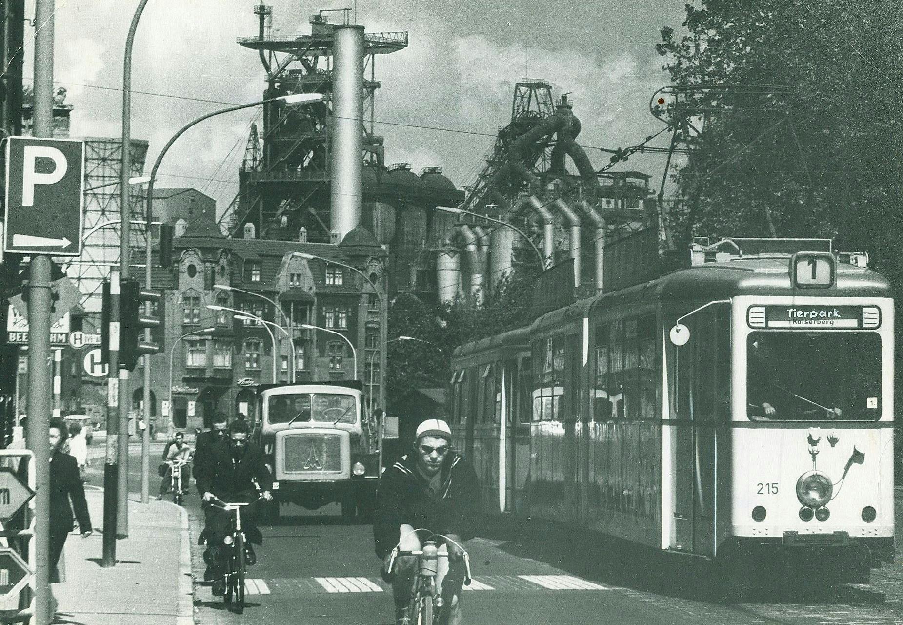 Großstädtische Straßenszene mit Straßenbahn, Lastwagen, Fahrradfahrern und Fußgängern. Das Bild ist so aufgenommen, dass alle auf einen zu kommen. Im Hintergrund das Stahlwerk Thyssen.